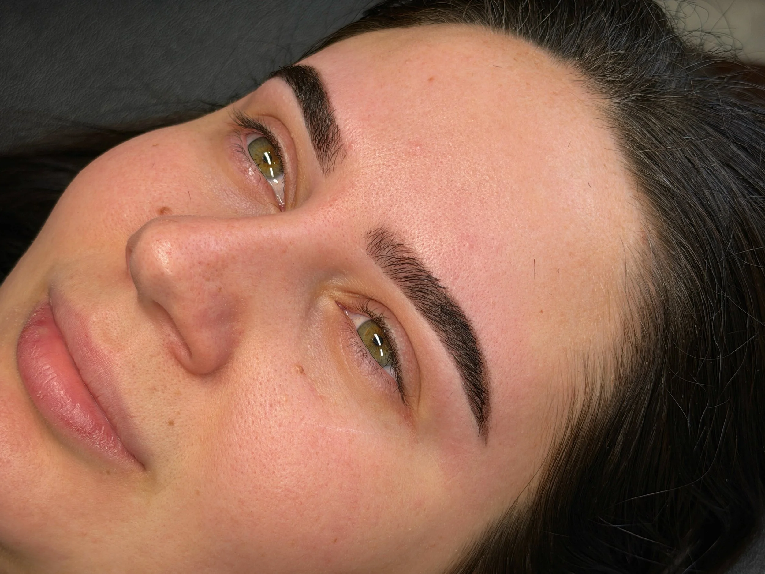 Close-up of a woman's face showing her with dark hair, well-groomed eyebrows, and light makeup, lying down.