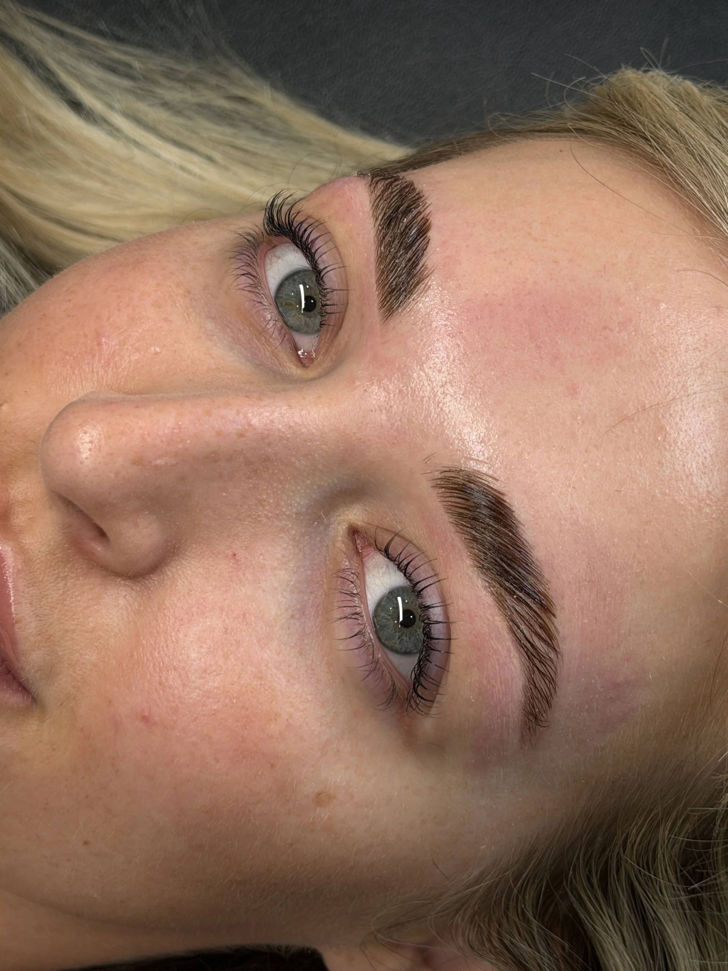 Close-up of a woman's face with freshly groomed eyebrows and long eyelashes, lying down with her hair visible in the background.