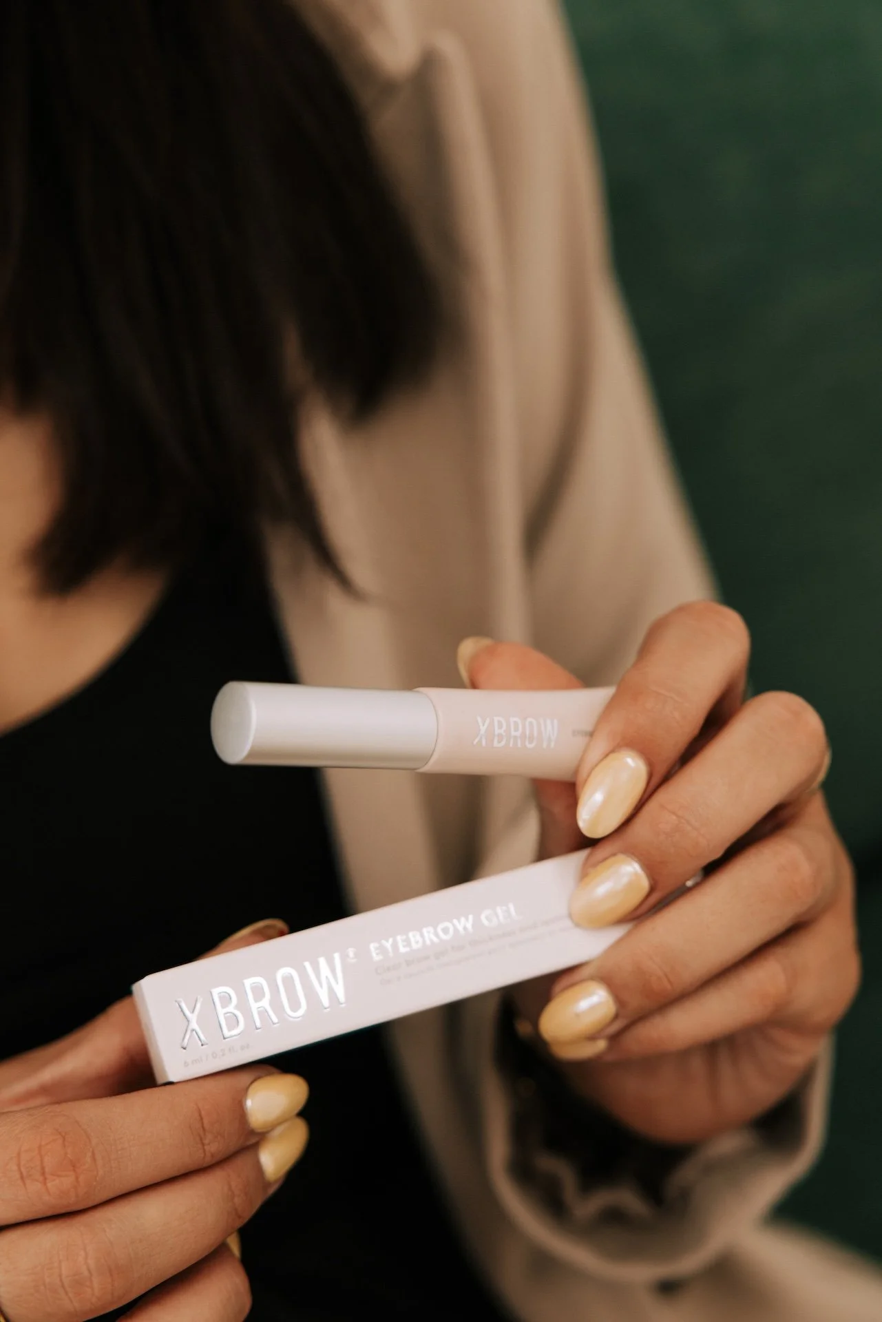 Close-up of a person holding a tube of XBROW eyebrow gel and its box, with manicured nails and part of their face partially visible.