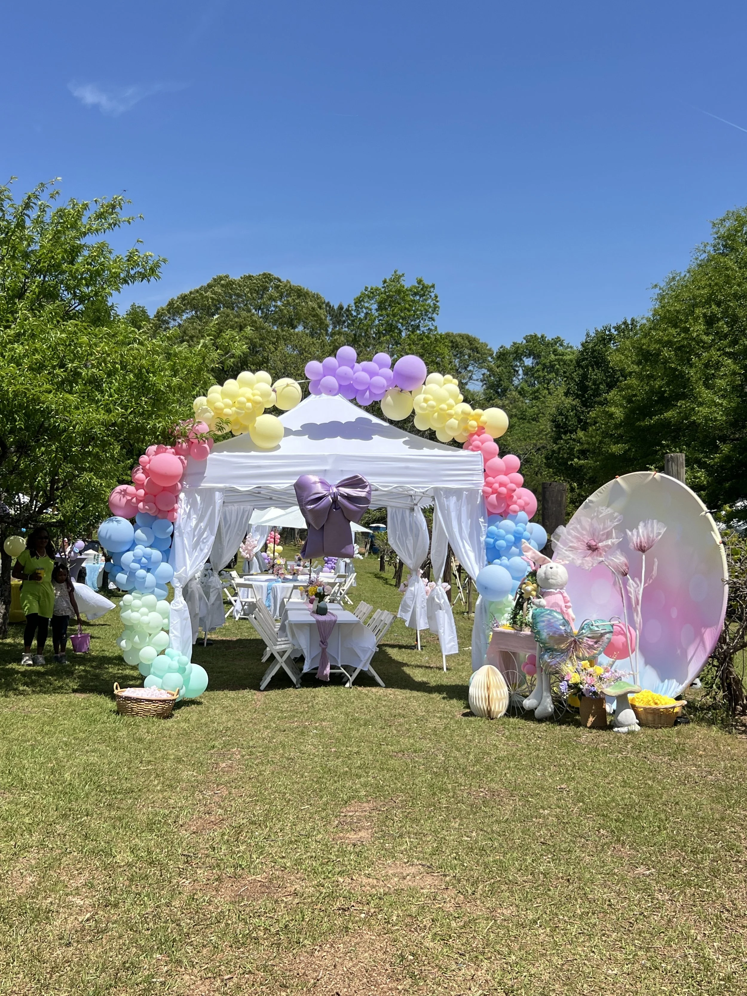 Decorated outdoor party scene with a white canopy tent, pastel-colored balloons, tables with pink and purple tablecloths, and children playing on the grass.