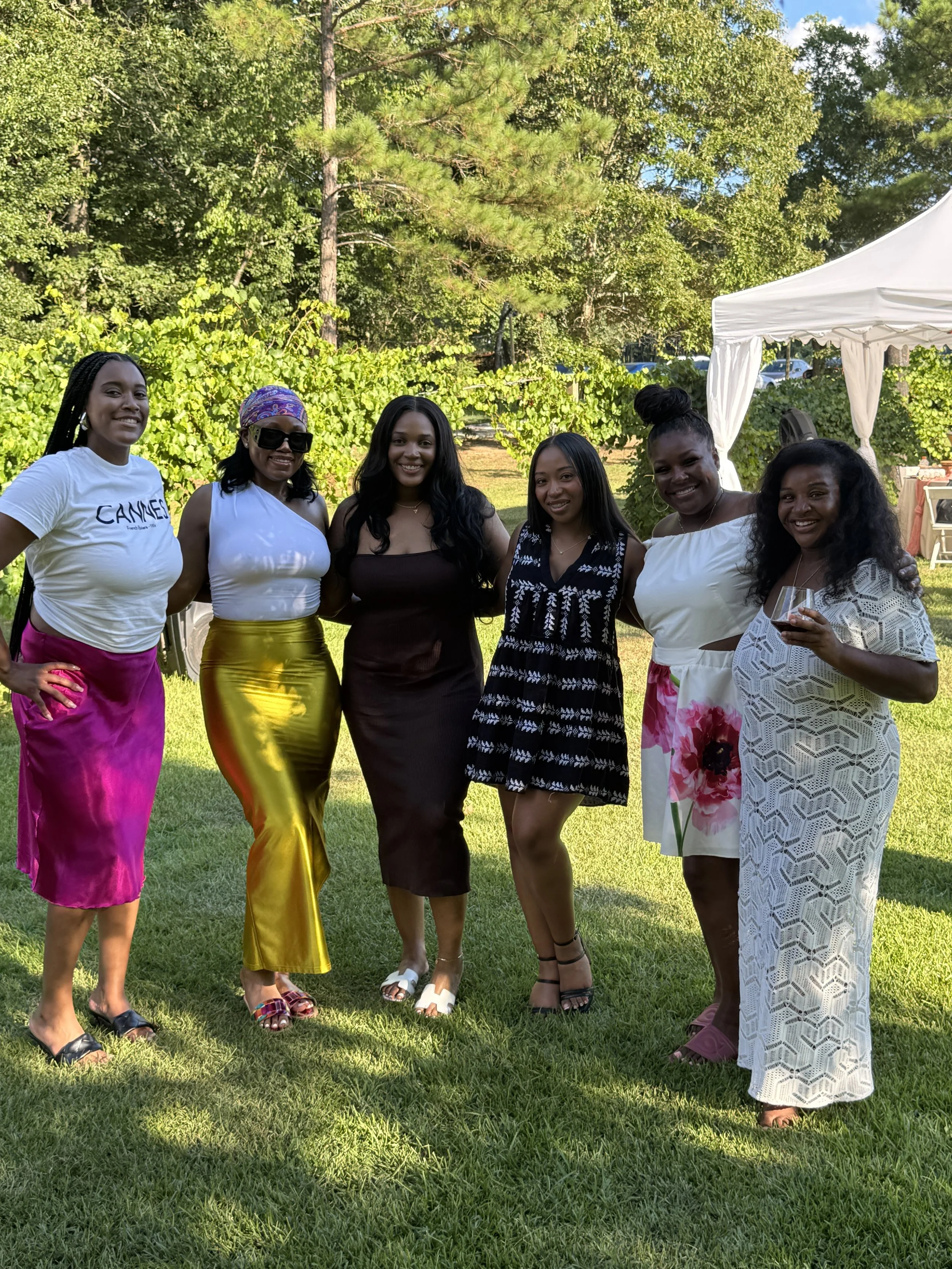 Group of seven women standing on grass outdoors, smiling, dressed in colorful summer outfits, with trees and a white canopy in the background.