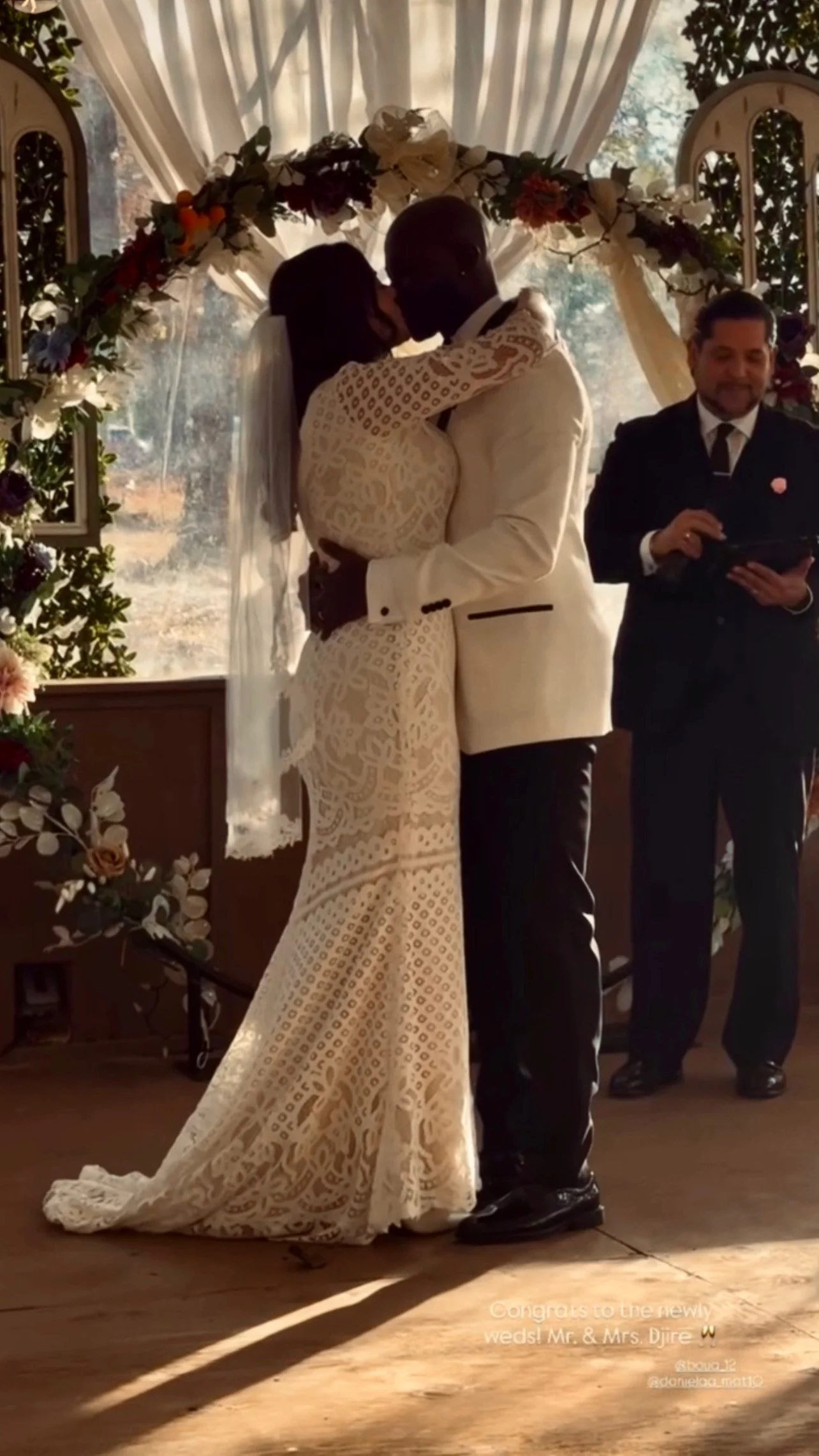 A bride and groom share a kiss during their wedding ceremony, standing beneath a decorated arch with flowers. An officiant or ring bearer stands nearby, holding a tablet and smiling.
