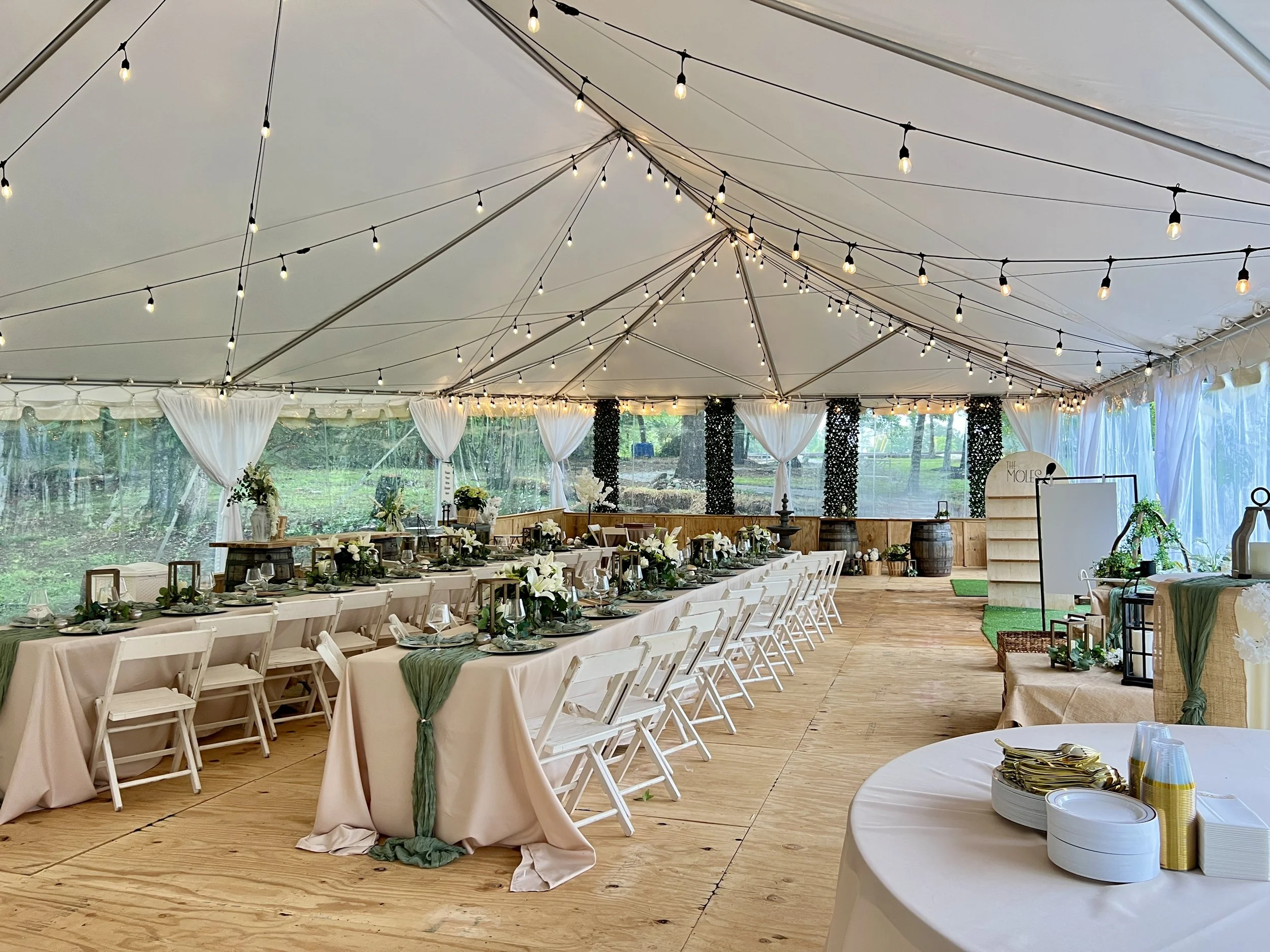 Decorated event tent interior with long dining tables, white chairs, floral centerpieces, string lights, and a refreshment or gift table.