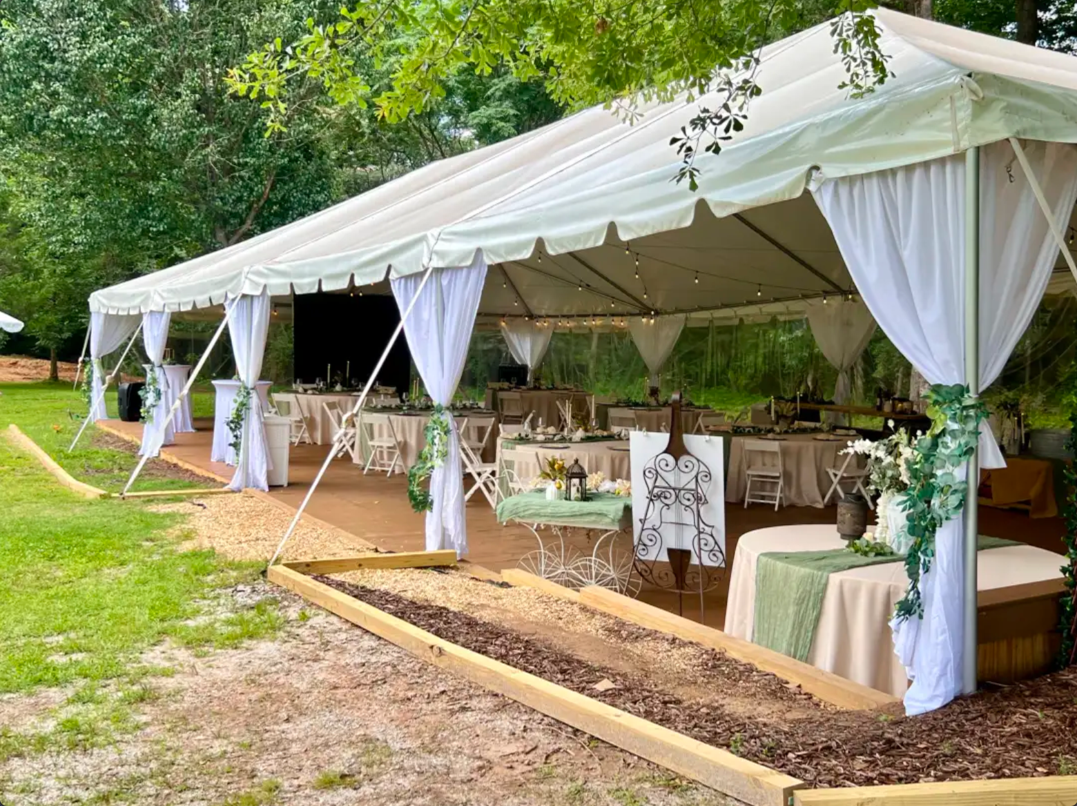 A large white outdoor tent decorated with lights and greenery, set up on a grassy area with trees, for a celebratory event or wedding reception.
