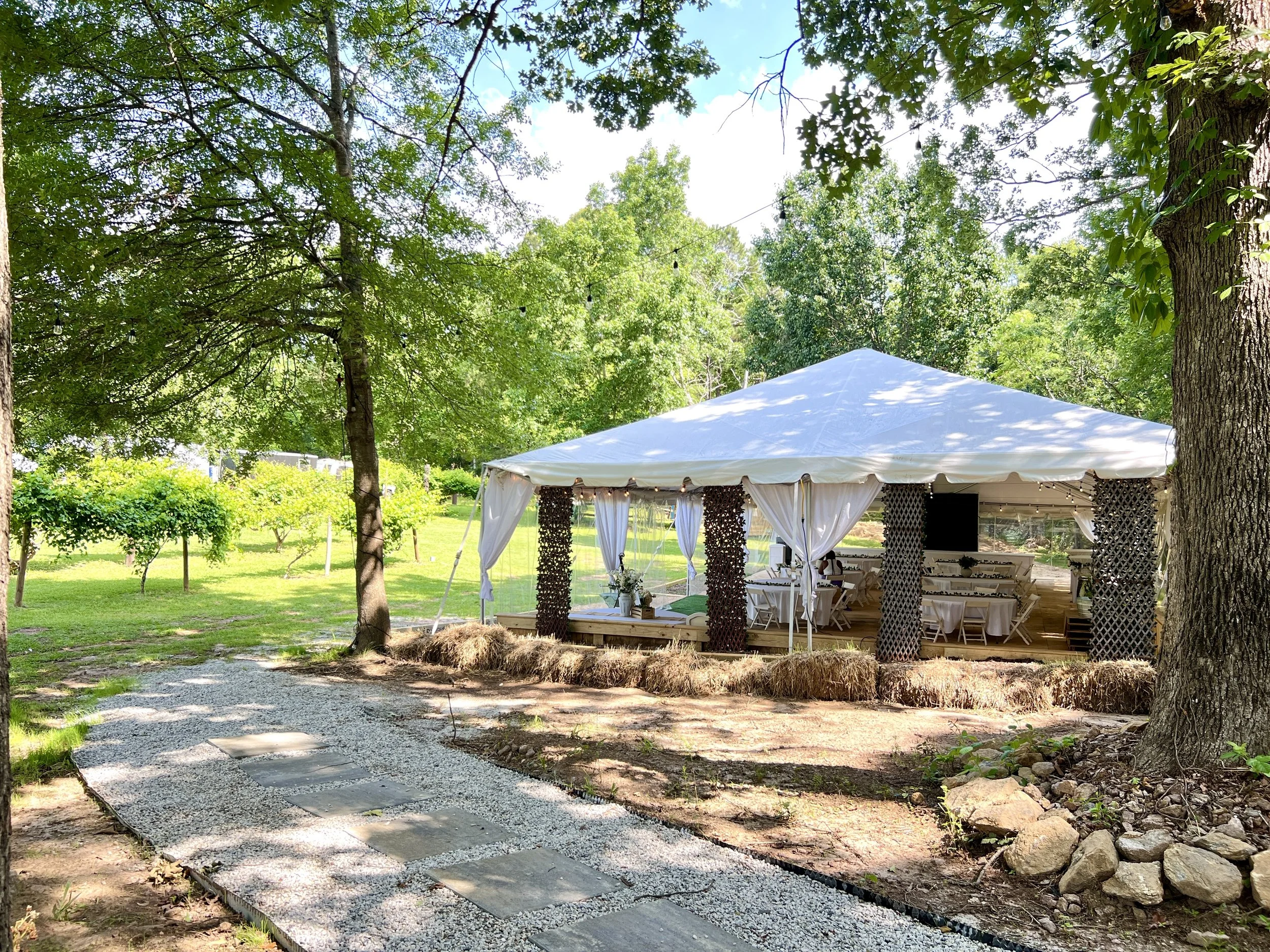 White event tent set up outdoors on a grassy area surrounded by trees with a stone pathway leading to it, decorated with string lights and surrounded by natural landscaping.