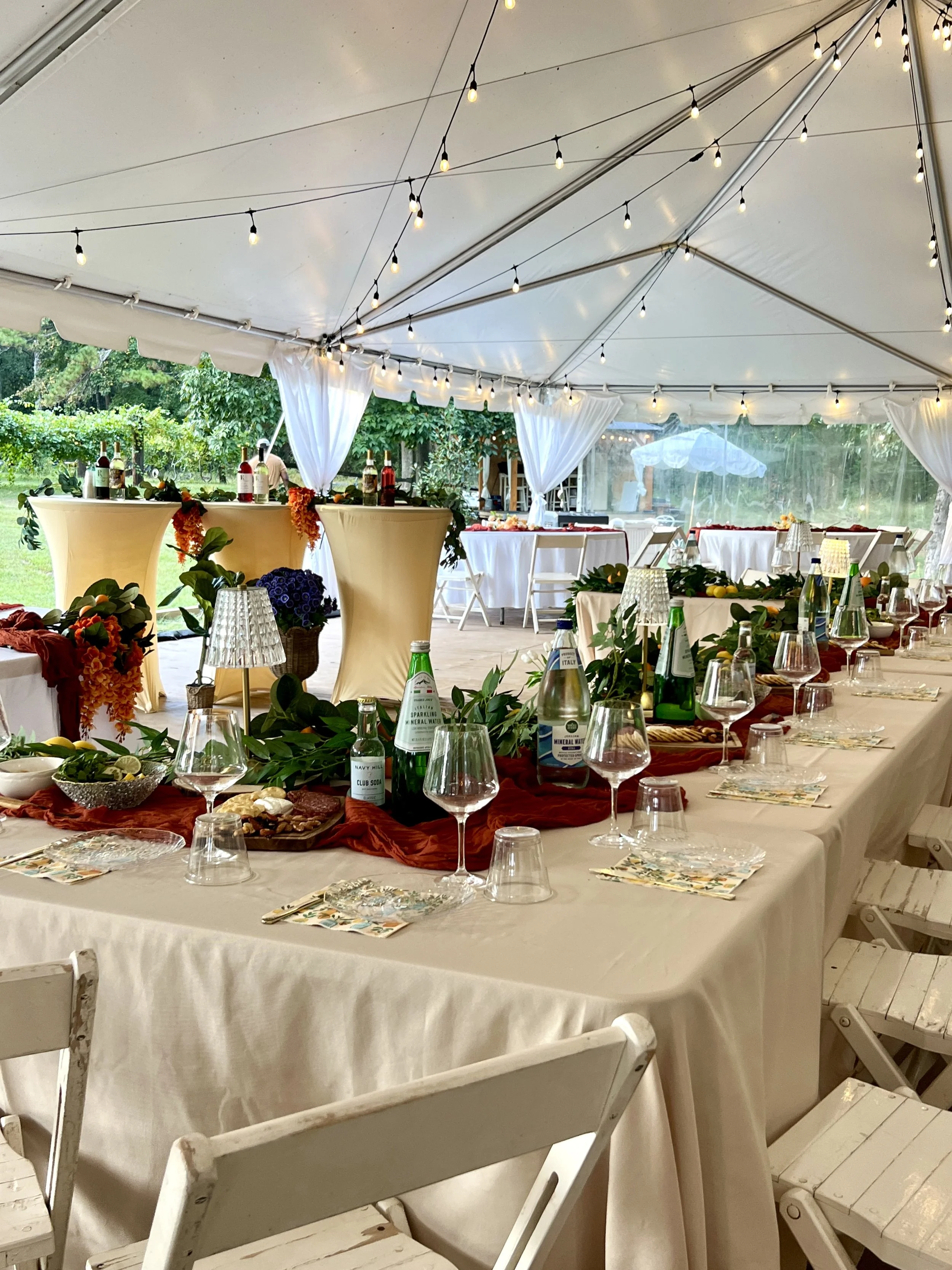 Decorated outdoor party tent with string lights, tables set with glasses and floral arrangements, and a scenic garden view.