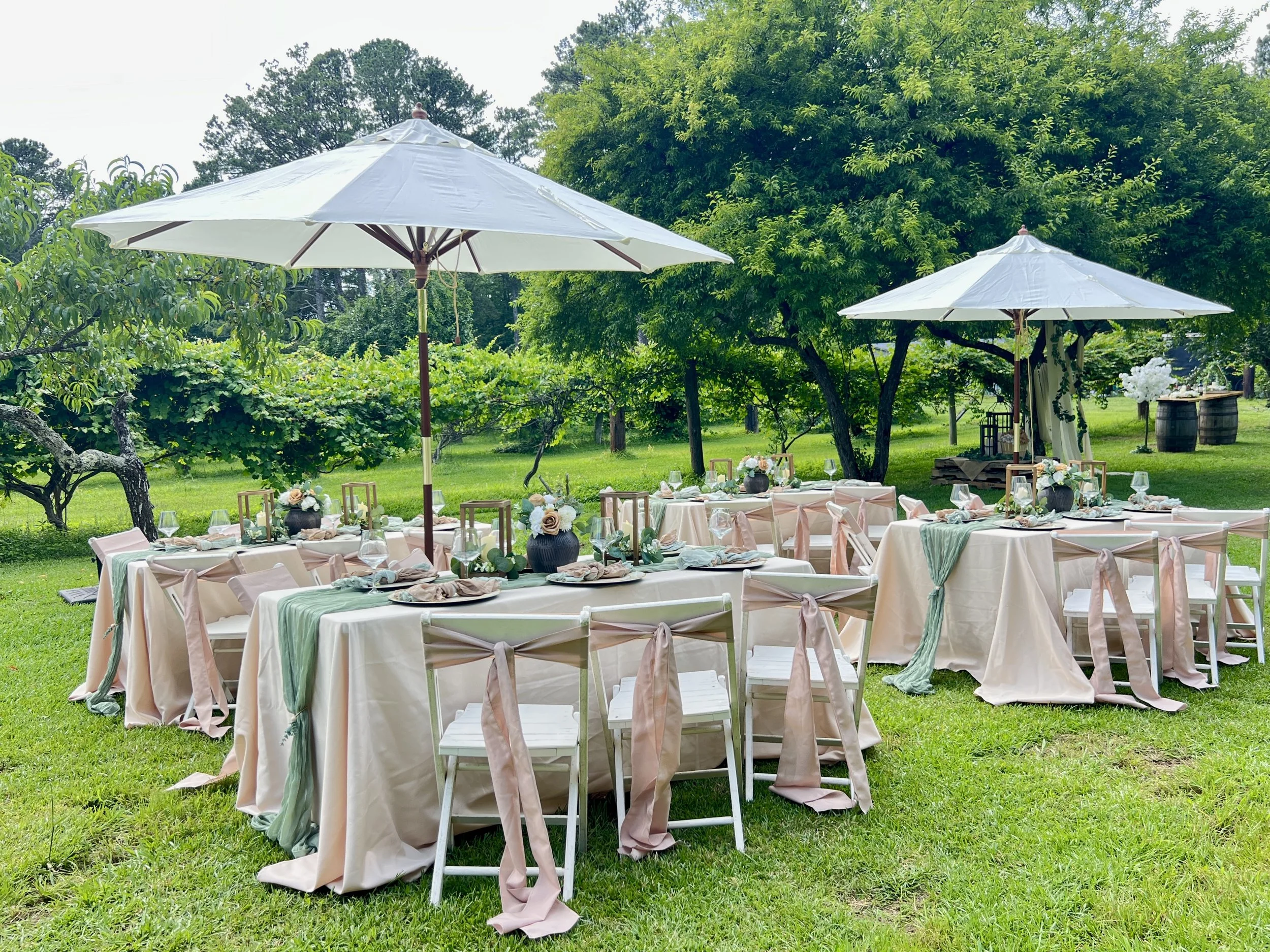 Outdoor event setup with decorated tables, chairs, umbrellas, and floral centerpieces in a lush green garden.
