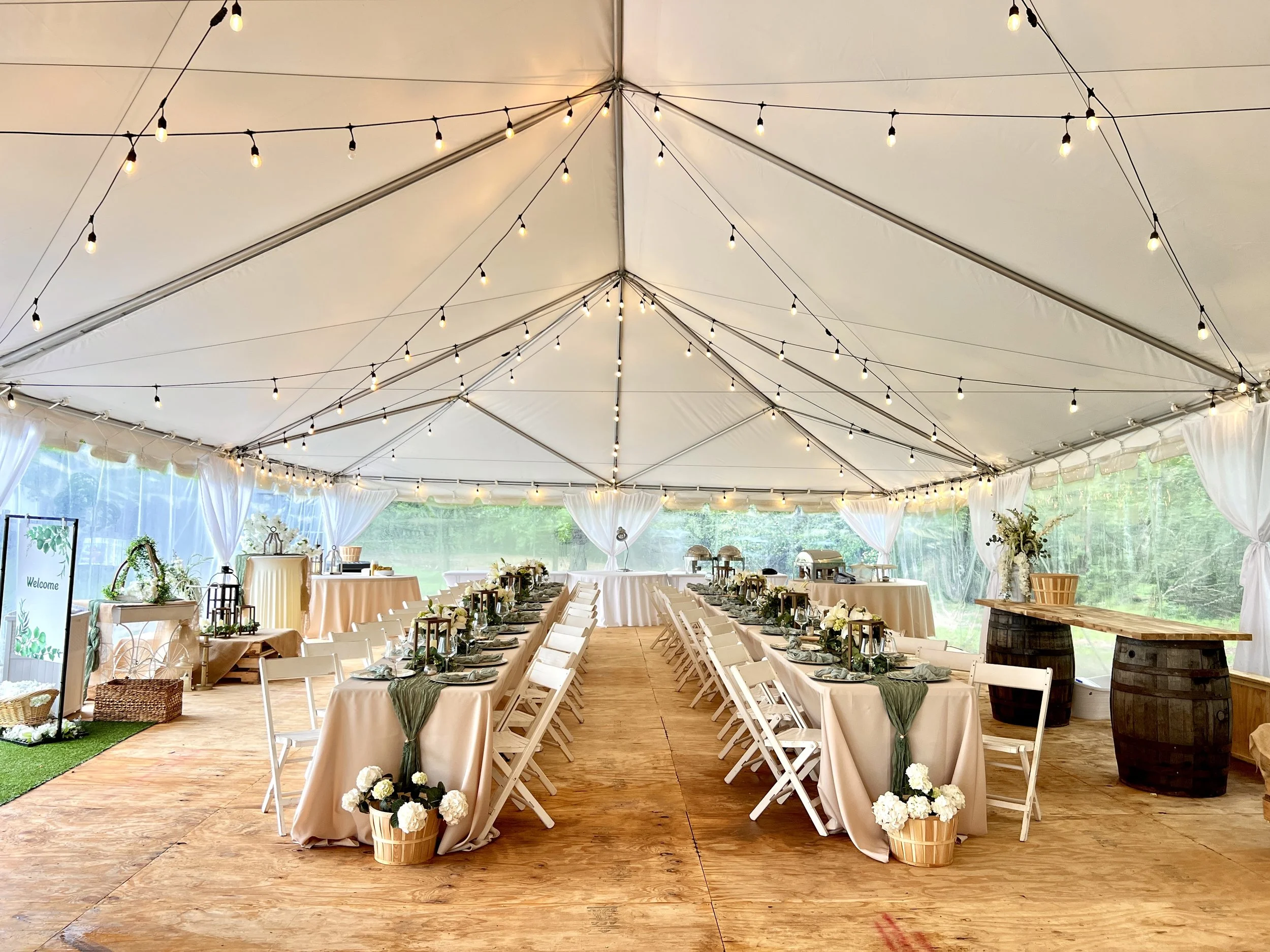 Outdoor wedding reception setup inside a large white tent with string lights, decorated tables with beige tablecloths, flowers, and place settings, and a wooden floor.