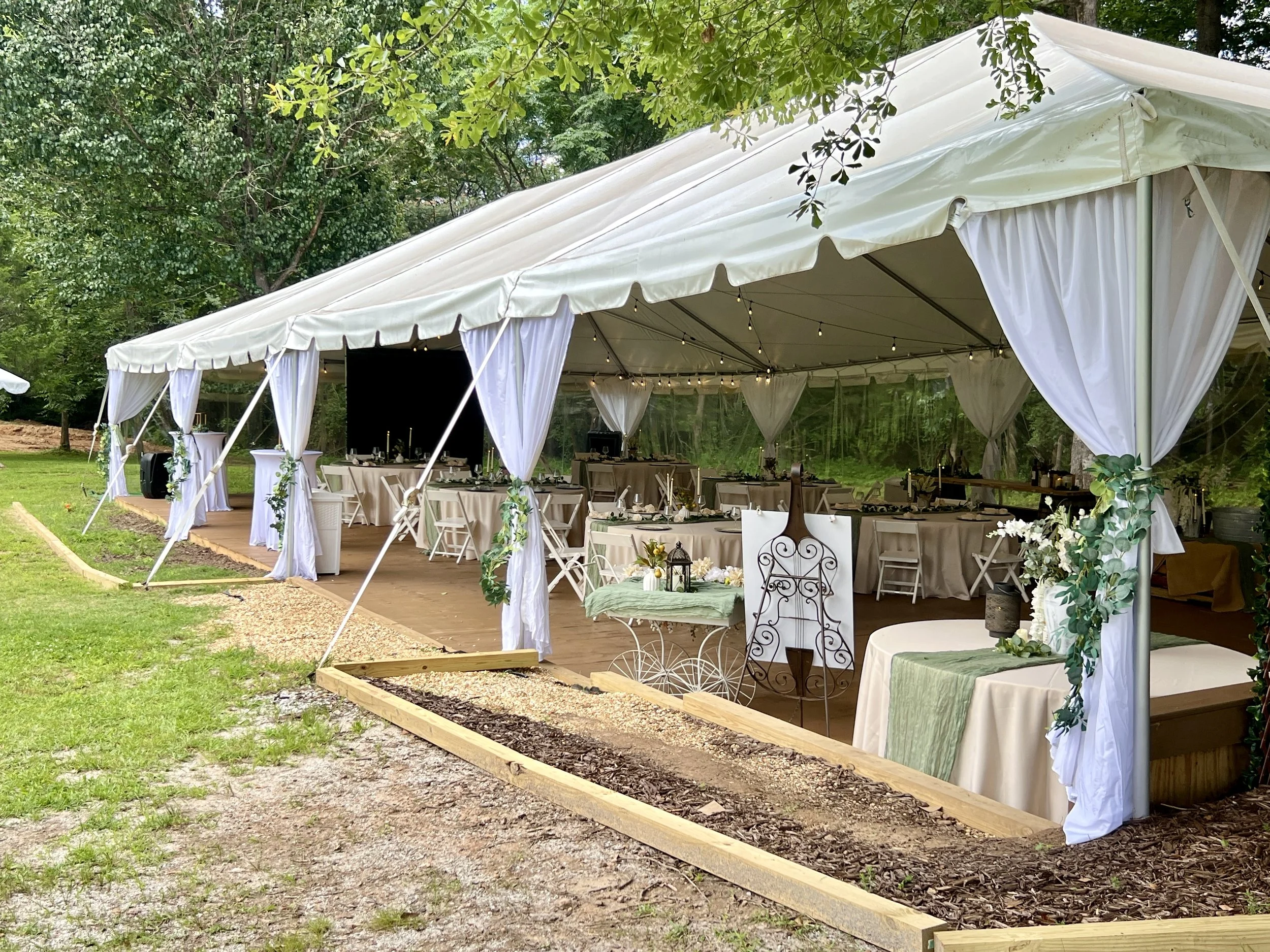 Decorated outdoor event tent with white curtains and tables set for a celebration, surrounded by green trees.