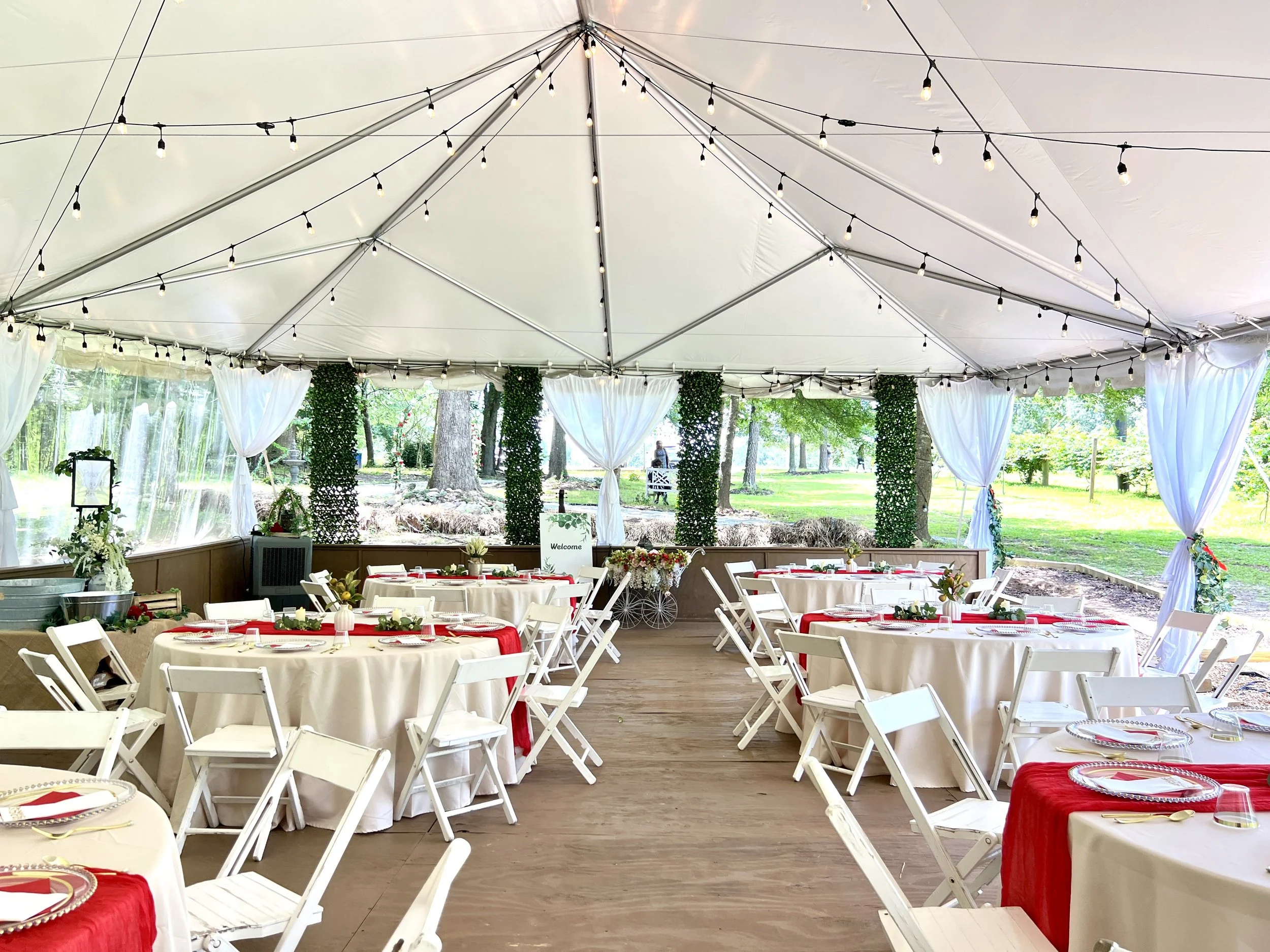 An outdoor wedding reception area with white folding chairs and tables covered with white tablecloths, set under a large tent with string lights and white curtains, surrounded by trees and greenery.