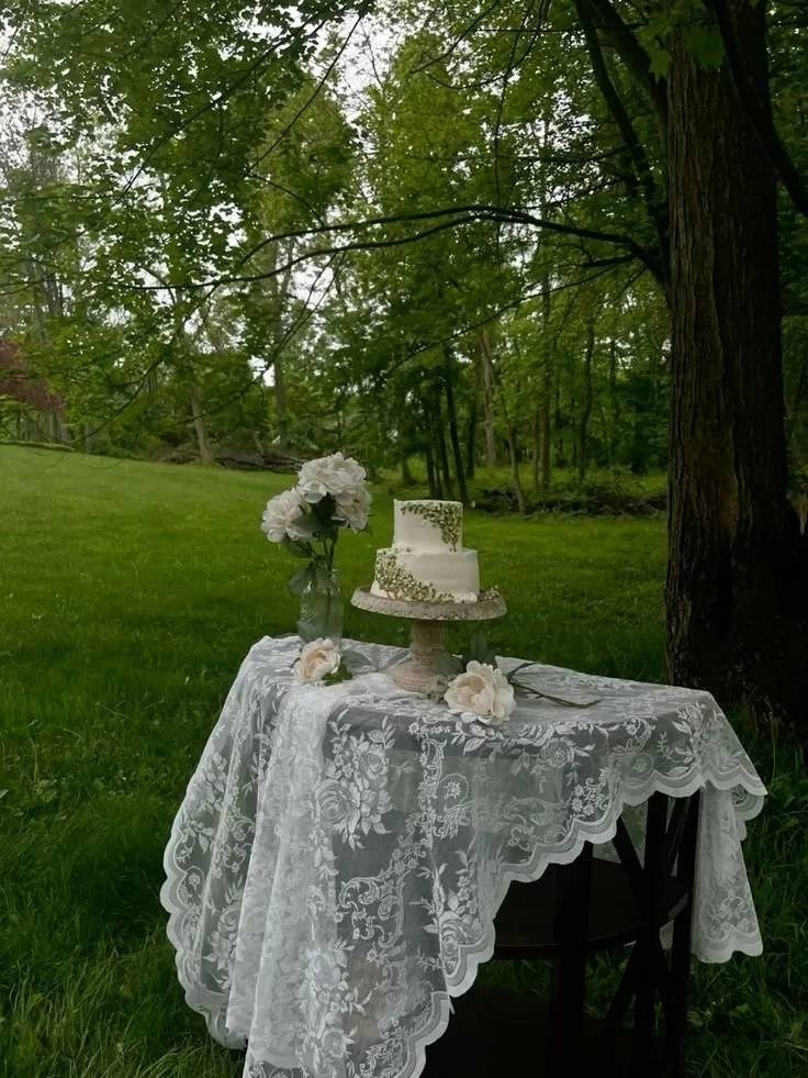 A white wedding cake with green decorations on a table with a lace tablecloth, set outdoors in a lush green area with trees.
