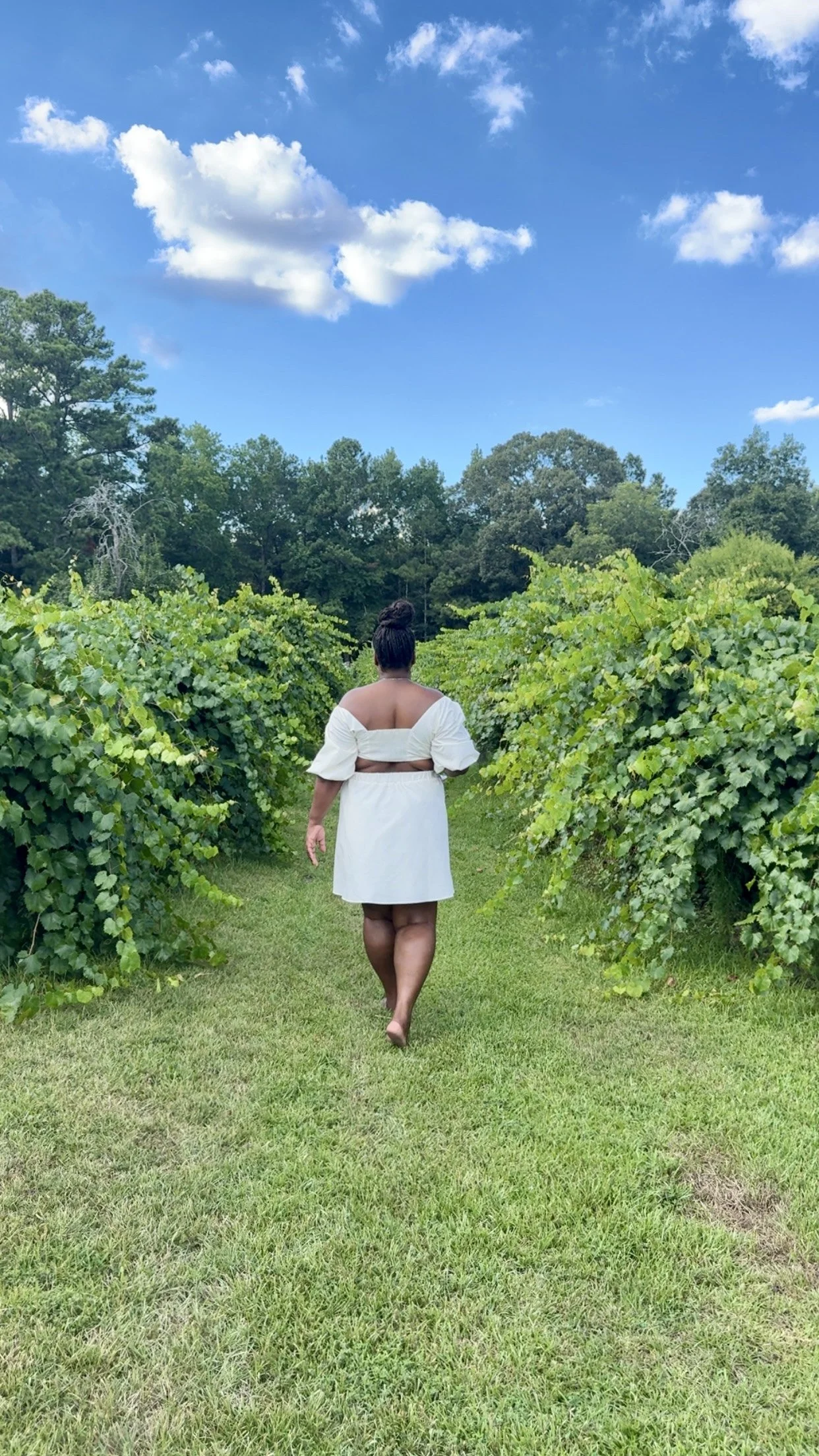 A woman walking barefoot through a vineyard or lush green field under a bright blue sky with a few clouds.