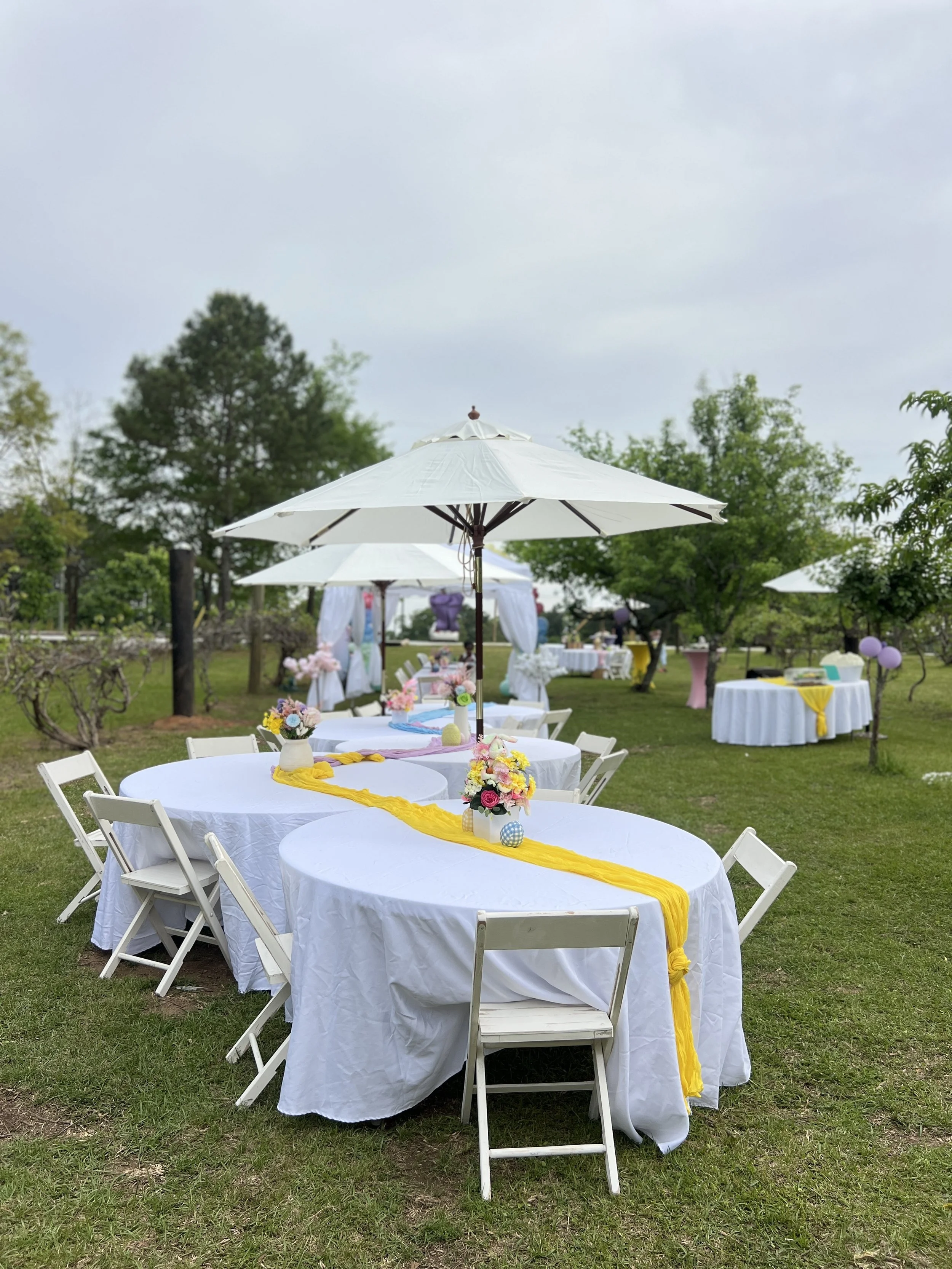 Decorated outdoor garden setup with round tables covered in white tablecloths, floral centerpieces, yellow runners, and white foldable chairs under large white umbrellas, likely for an event or celebration.