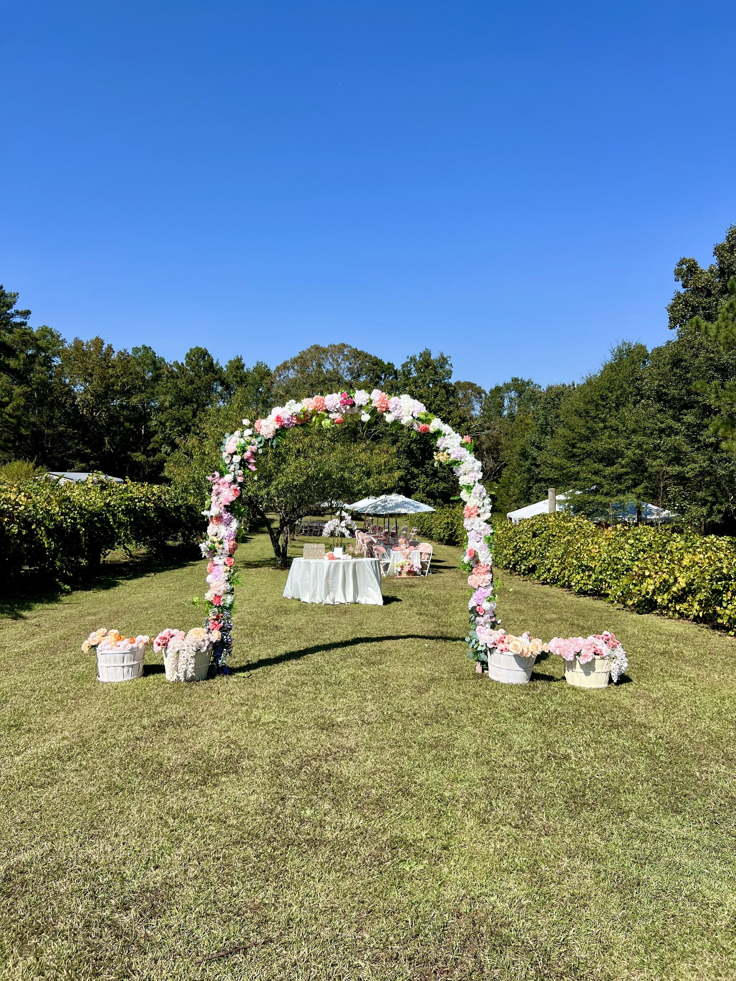 Decorated floral archway on grassy lawn with outdoor tables and chairs under umbrellas, surrounded by trees.