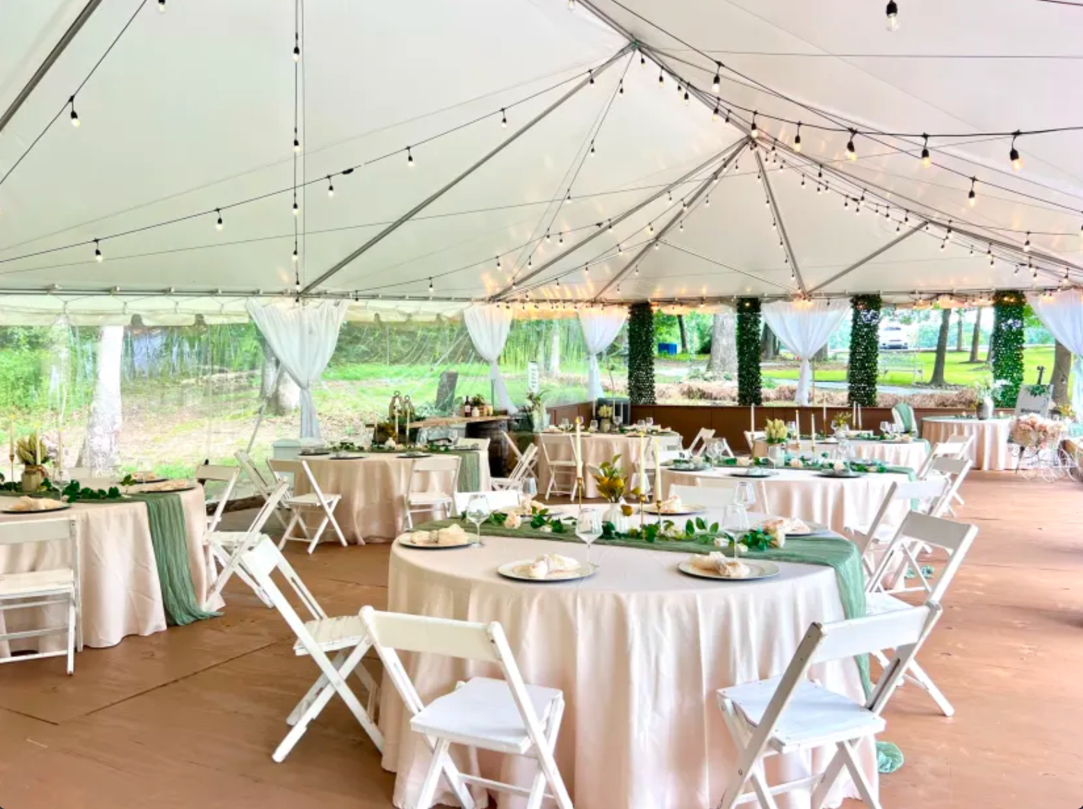 Decorated event tent interior with round tables covered in white tablecloths, green and white centerpieces, white chairs, string lights above, and greenery on the walls, set up for a celebration or wedding.