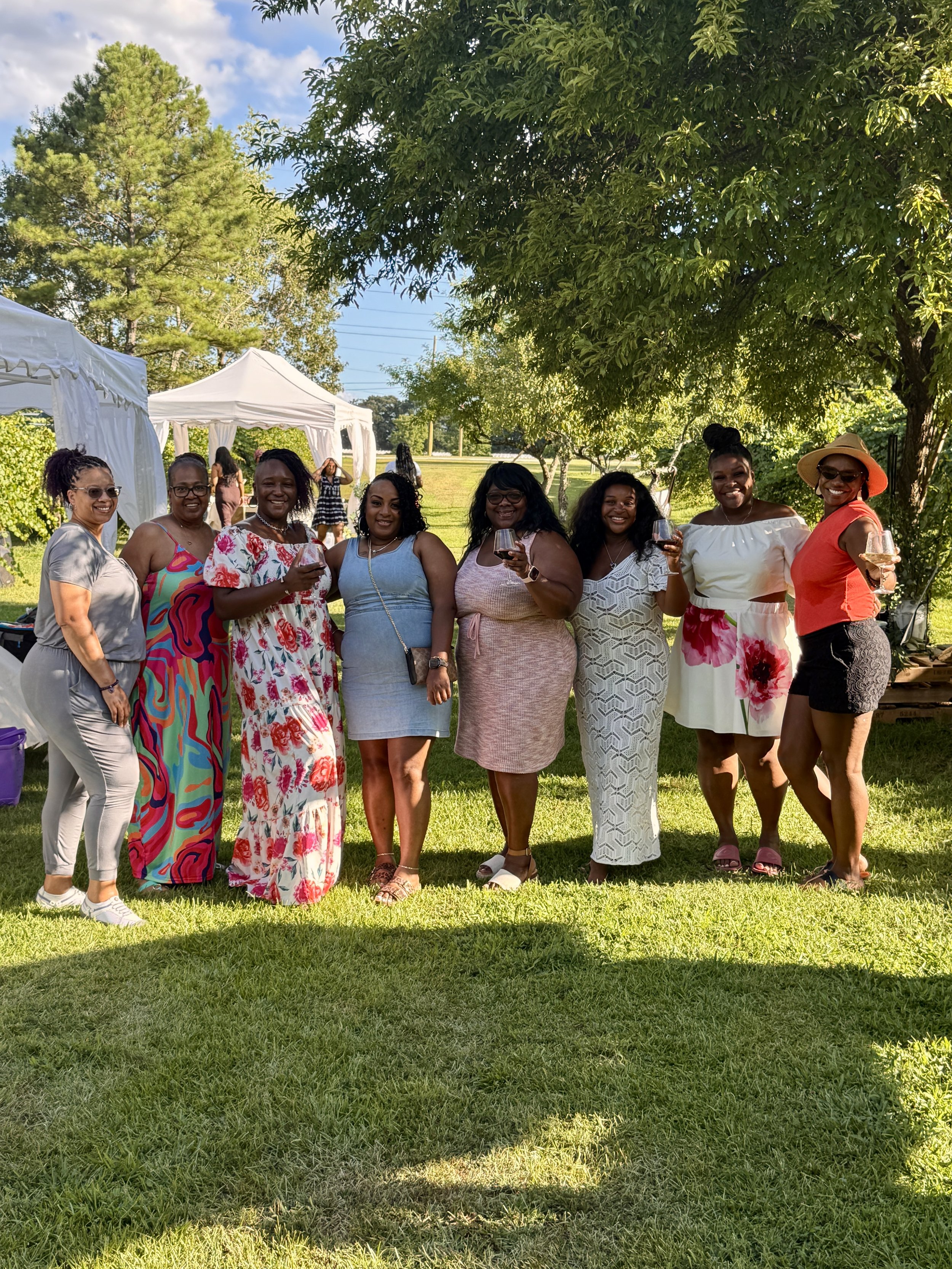 A group of nine women standing outdoors on grass, holding glasses of wine, smiling, in a sunny garden area with trees and white tents in the background.