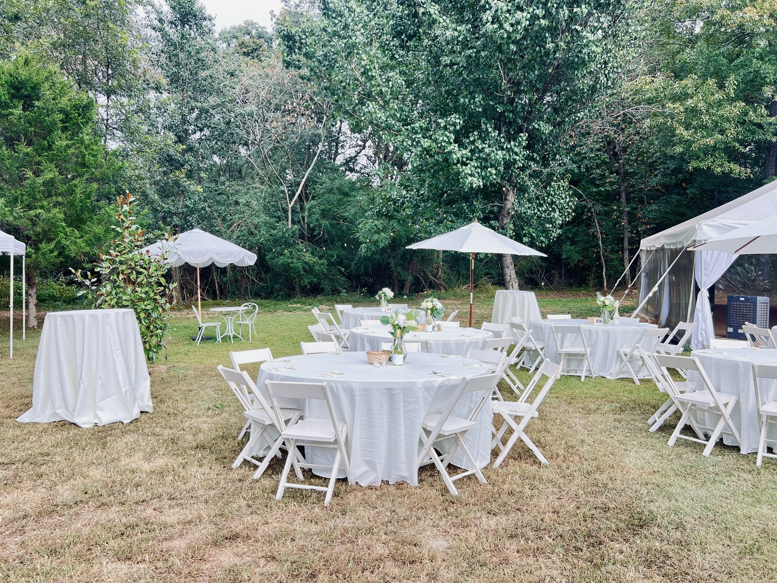 Outdoor event setup with round tables covered in white tablecloths, white chairs, white umbrellas, and greenery in the background.