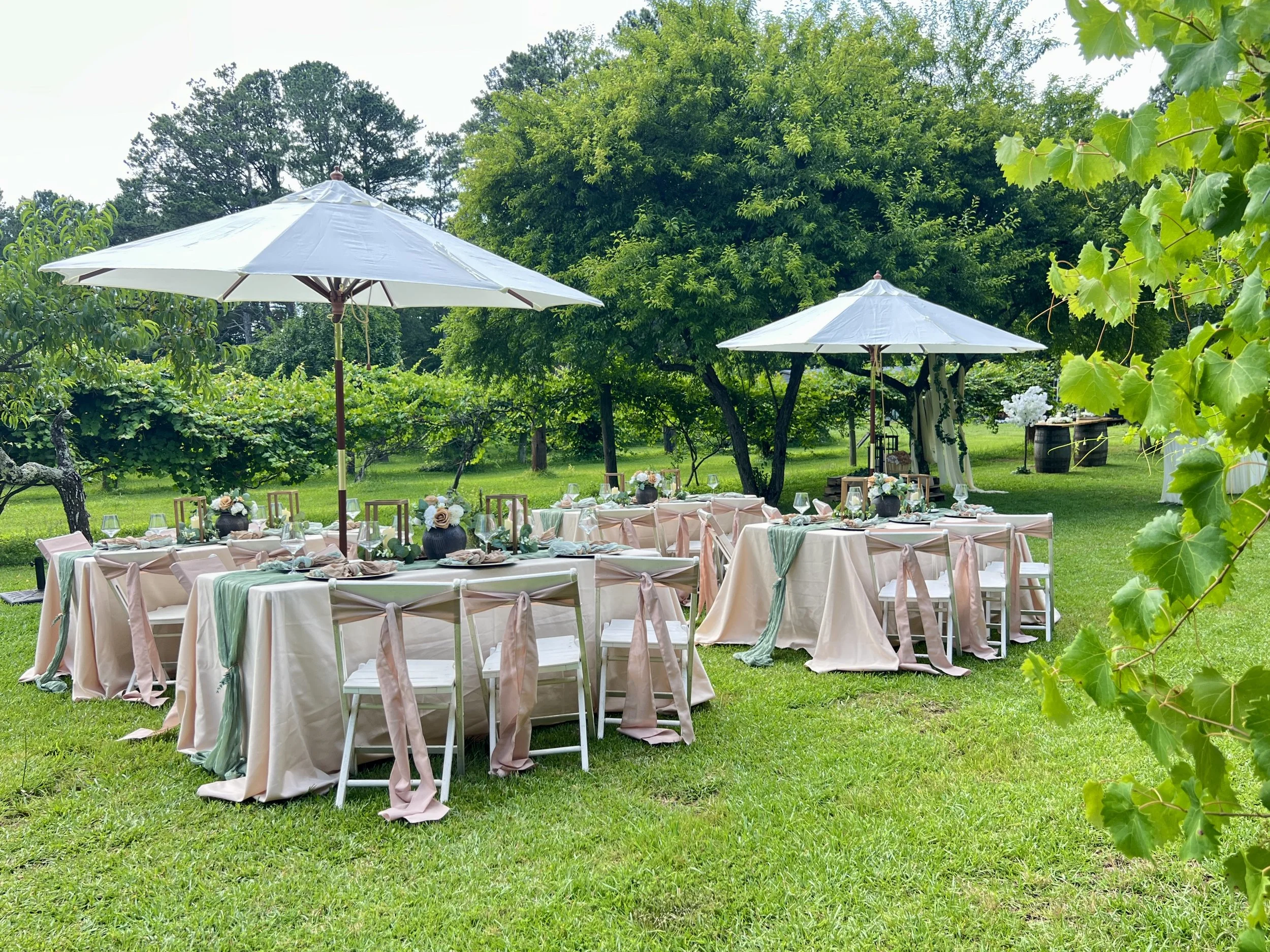 Outdoor garden party setup with round tables covered with pastel tablecloths, decorated with floral centerpieces, and surrounded by chairs with ribbons. Large white umbrellas provide shade on a grassy lawn with trees in the background.