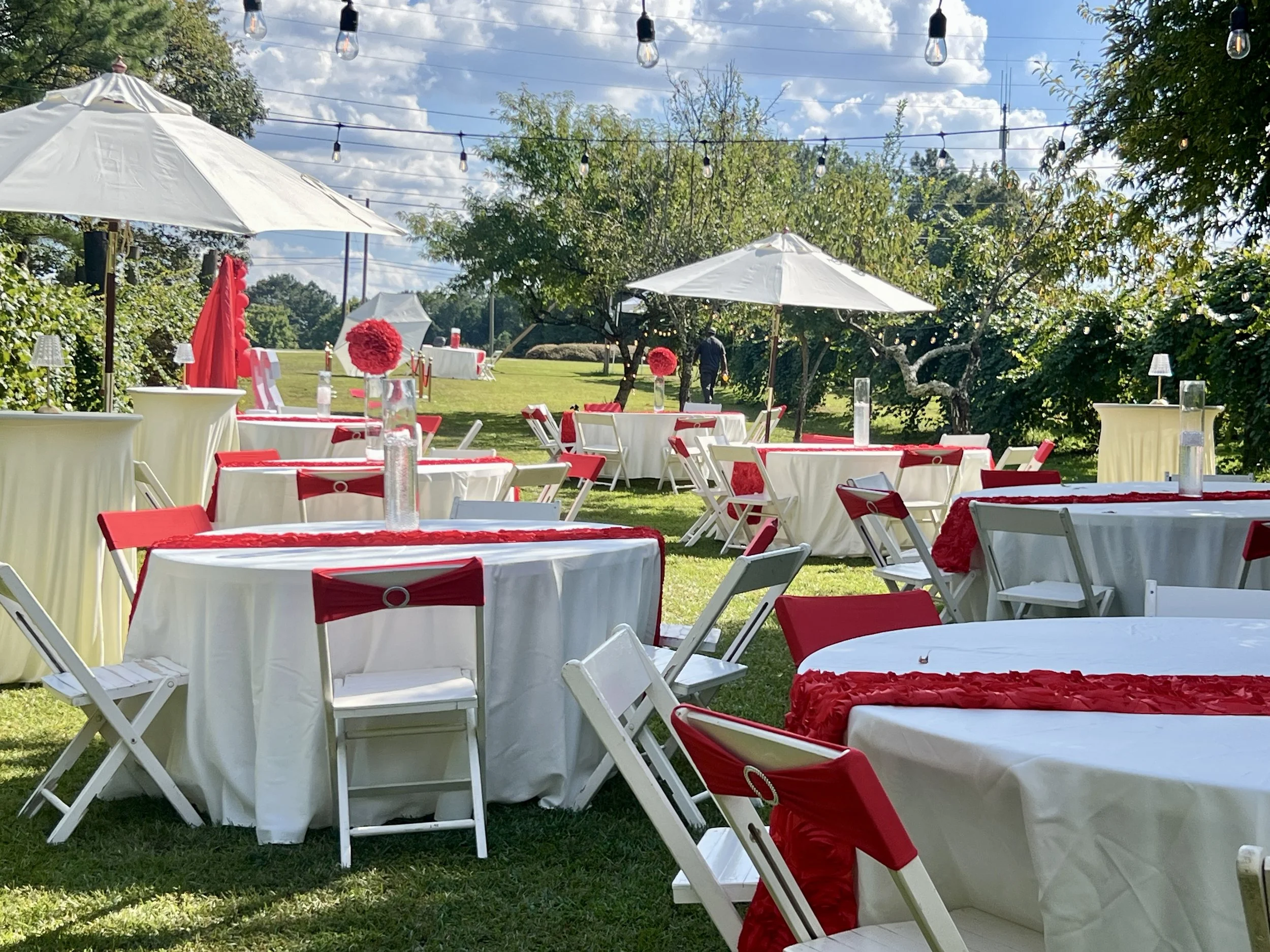 Outdoor event setup with round tables covered in white tablecloths, decorated with red accents, surrounded by white chairs with red sashes, under large white umbrellas, with string lights overhead, on a grassy area with trees and a partly cloudy sky.