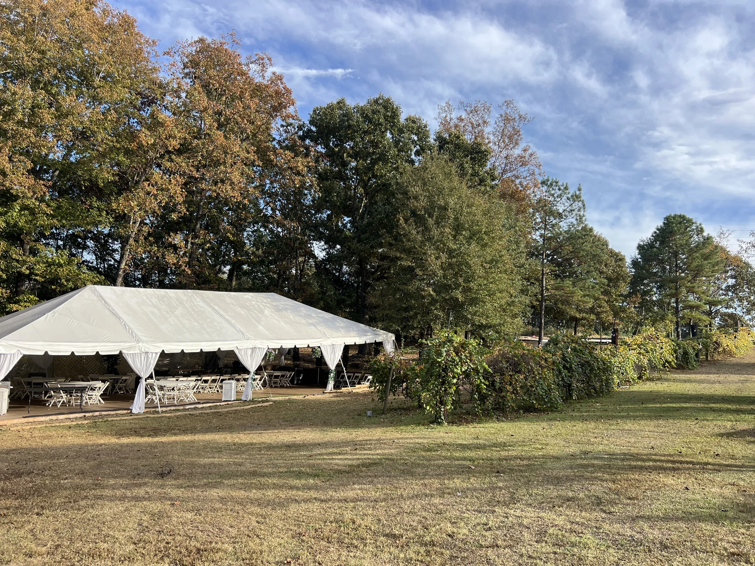 A large white event tent set up outdoors on a grassy area, with trees in fall foliage and a bright blue sky with some clouds in the background.