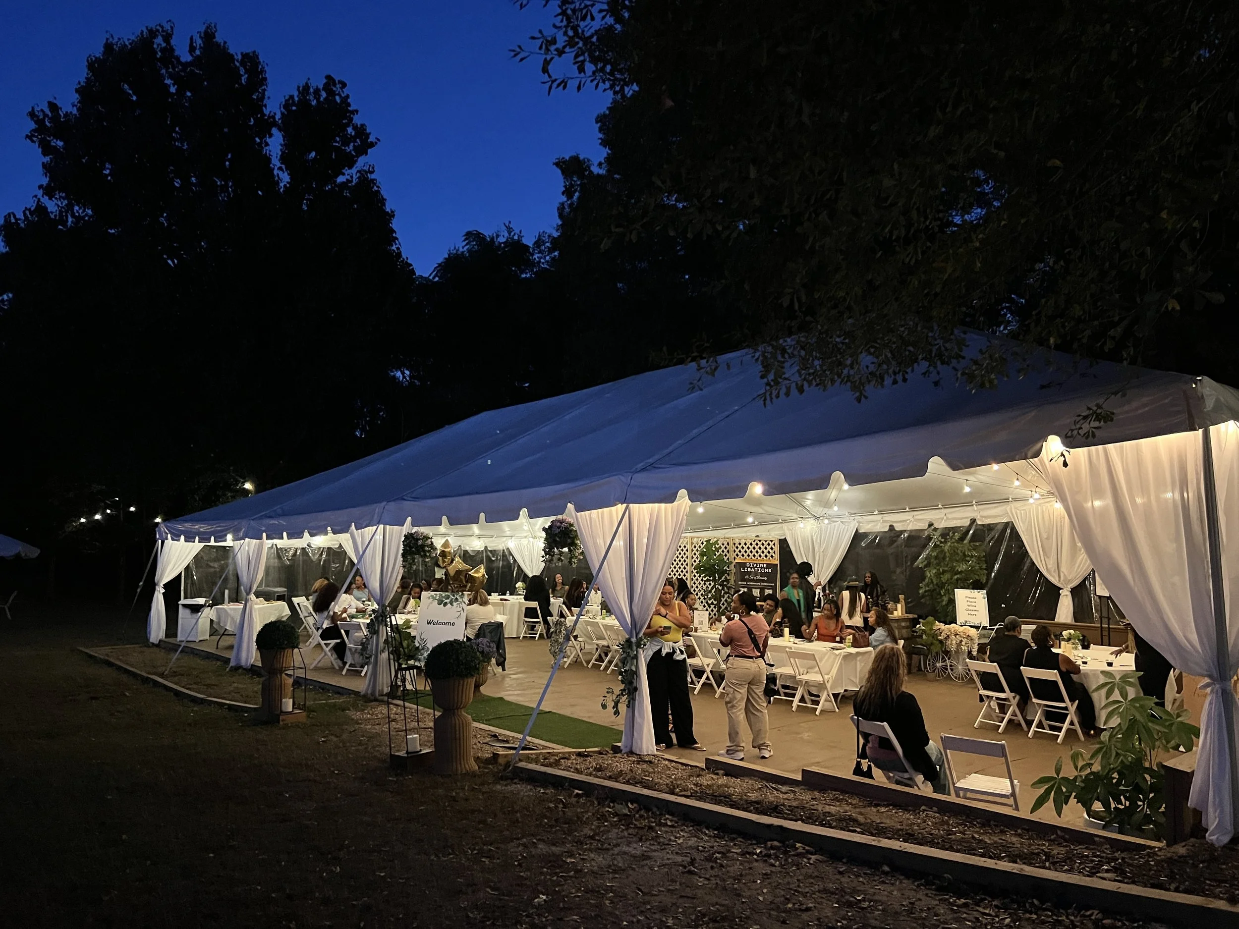 A nighttime outdoor celebration under a large white tent with string lights, decorated with flowers and greenery, with people sitting and standing around tables.