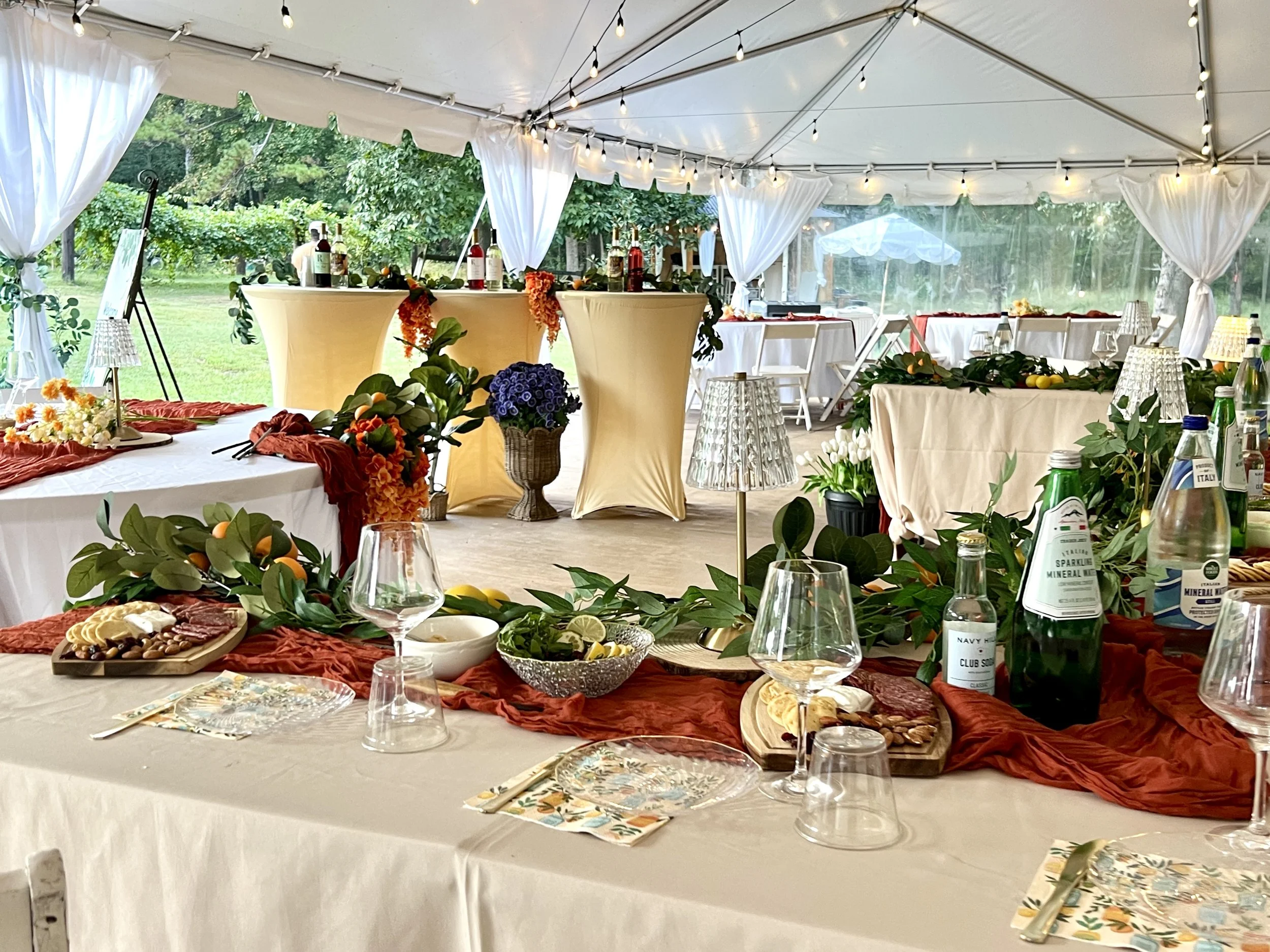 Decorated outdoor event tent with tables set for a celebration, featuring floral arrangements, wine bottles, and tableware, in a lush green garden setting.