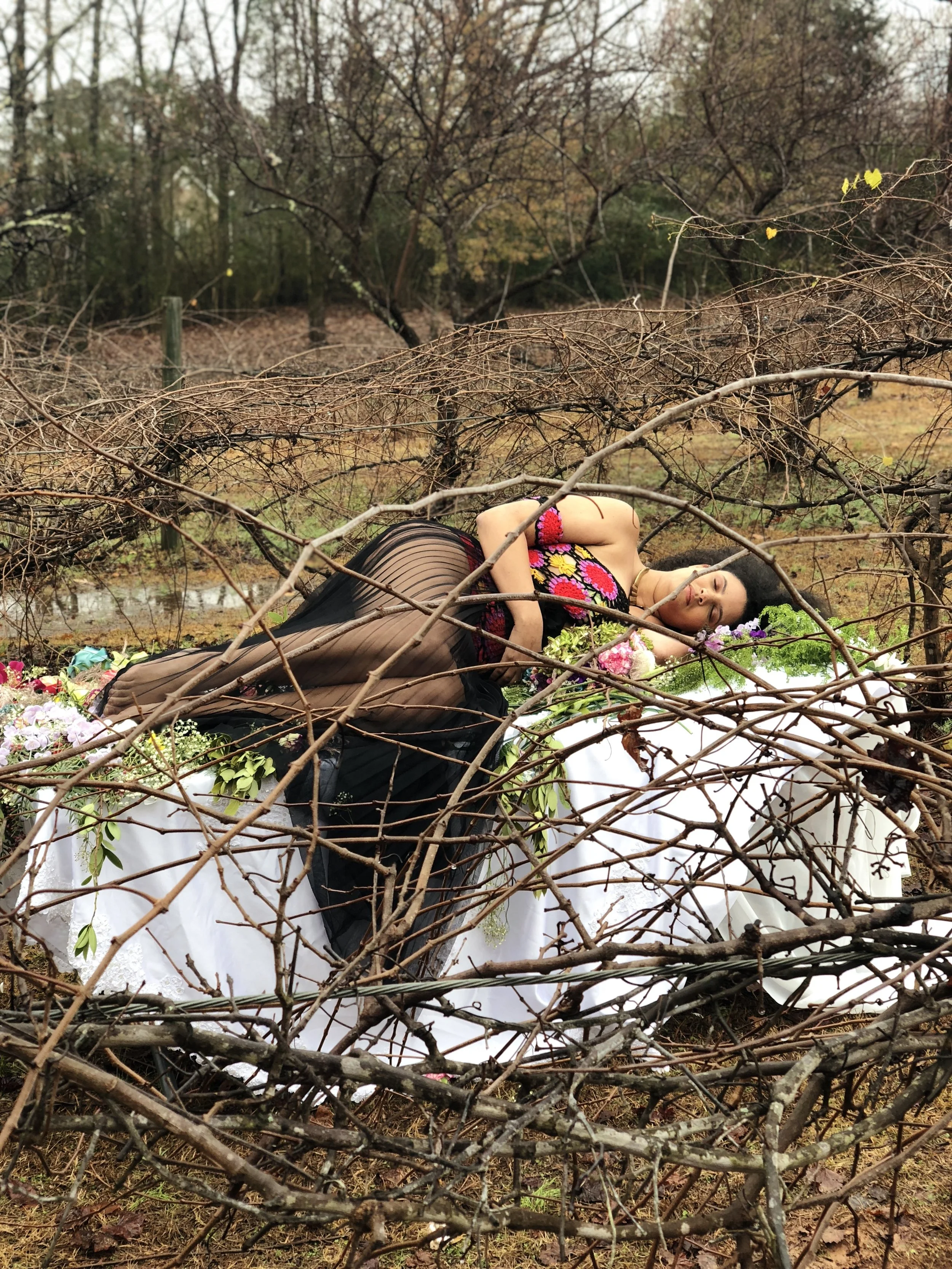 A woman lying on a table outdoors surrounded by leafless branches, with a floral dress and black stockings, seemingly posing in a natural setting.