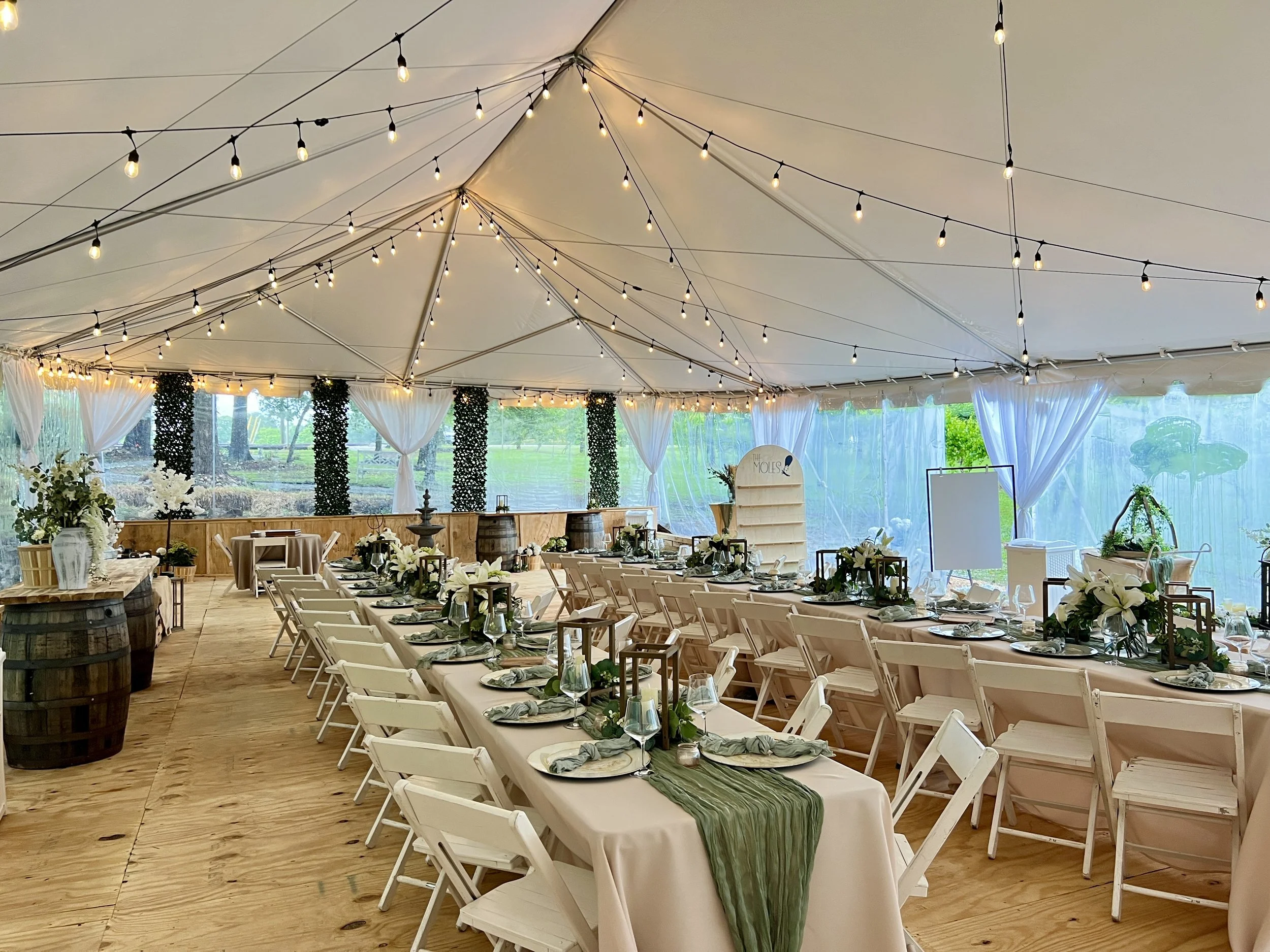 Event tent decorated for a celebration with long tables, white chairs, floral centerpieces, string lights, and a view of greenery outside.