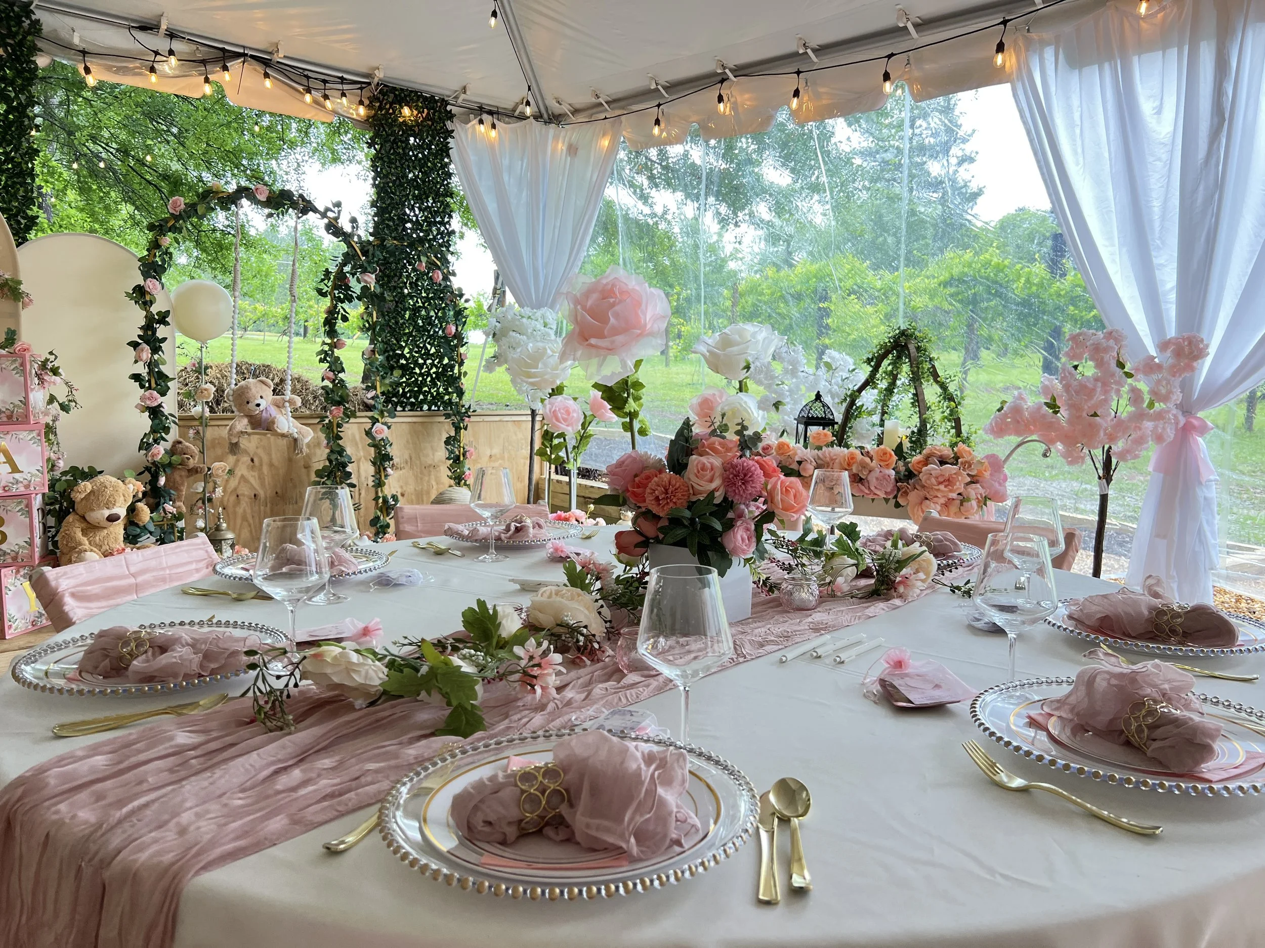 Elegant outdoor table set for a celebration with pink and white floral arrangements, teddy bears, hanging lights, and a swing decorated with flowers inside a white tent with white curtains and a view of a green garden outside.