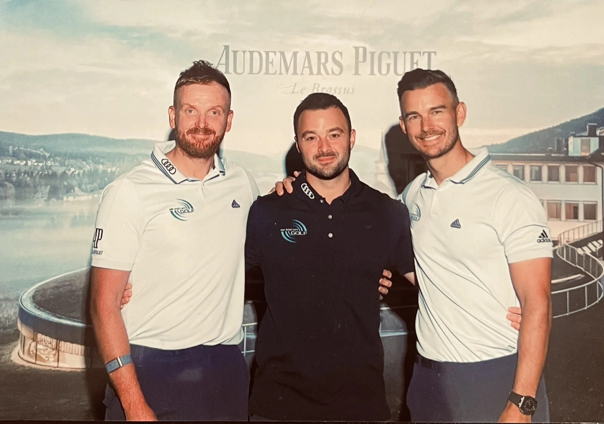 Three men smiling, two wearing white golf shirts with logos and one in a black shirt, standing together indoors against a backdrop showing the Audemars Piguet logo and an outdoor scene with mountains and a building.