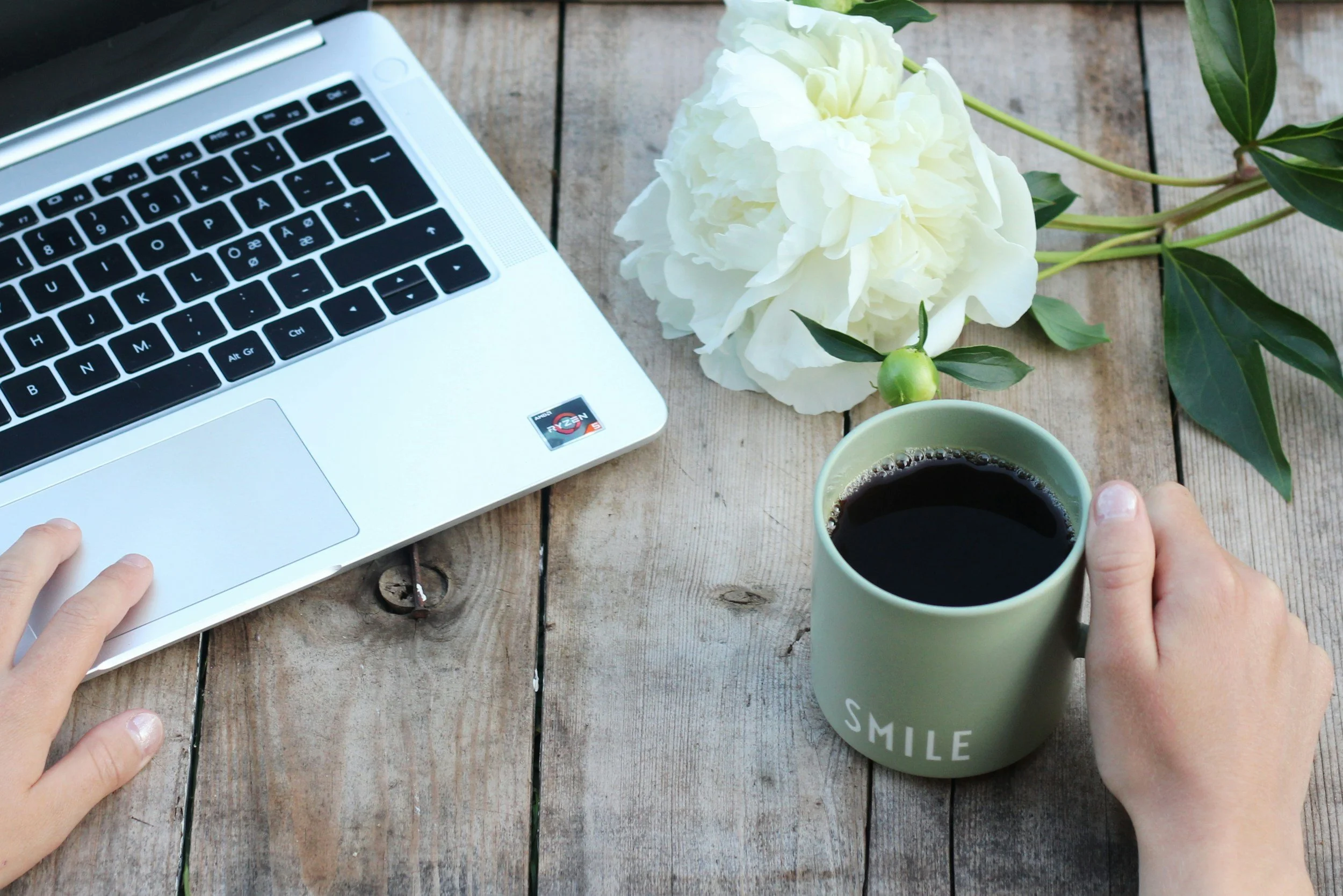 A person working on a laptop with a cup of coffee, a white flower, and green leaves on a wooden table.