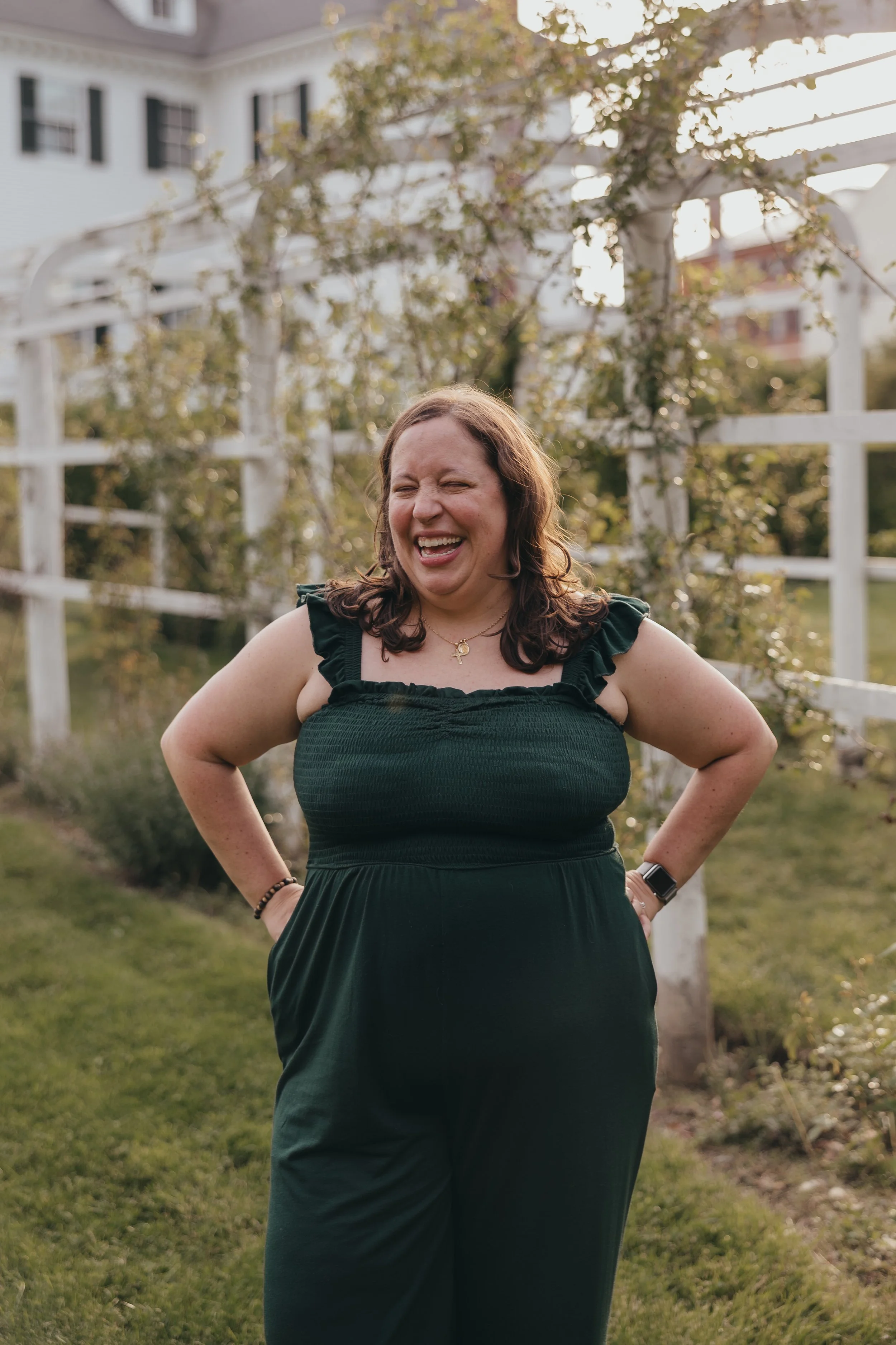 A photo of Amy Matos, bilingual storyteller for hire. She is in a dark green dress laughing outdoors in a garden with a white fence and greenery.
