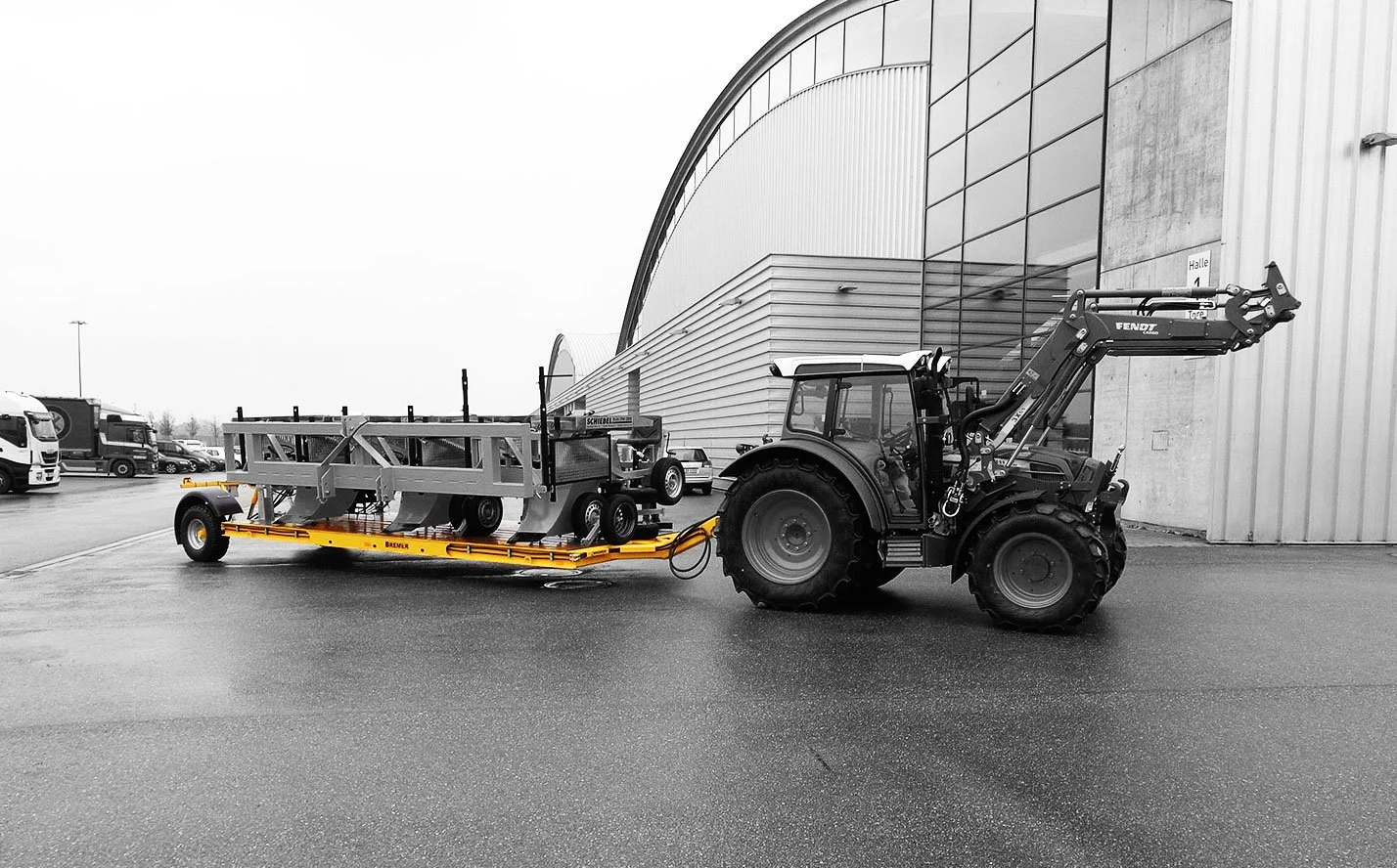 Ein Traktor zieht ein spezielles Transportfahrzeug, vermutlich für Baumaschinen oder schwere Geräte, vor einer modernen Industriehalle auf einer regennassen Asphaltfläche.