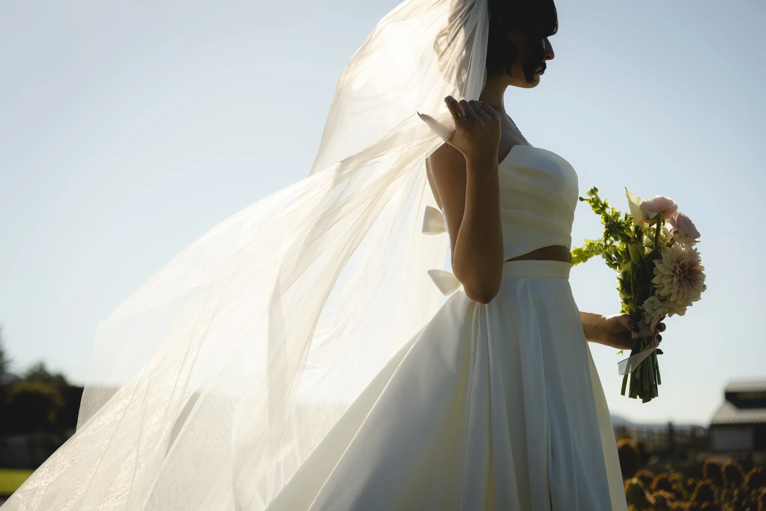 A woman holding a bouquet of flowers, wearing a wedding dress and veil, standing outdoors with sunlight behind her.