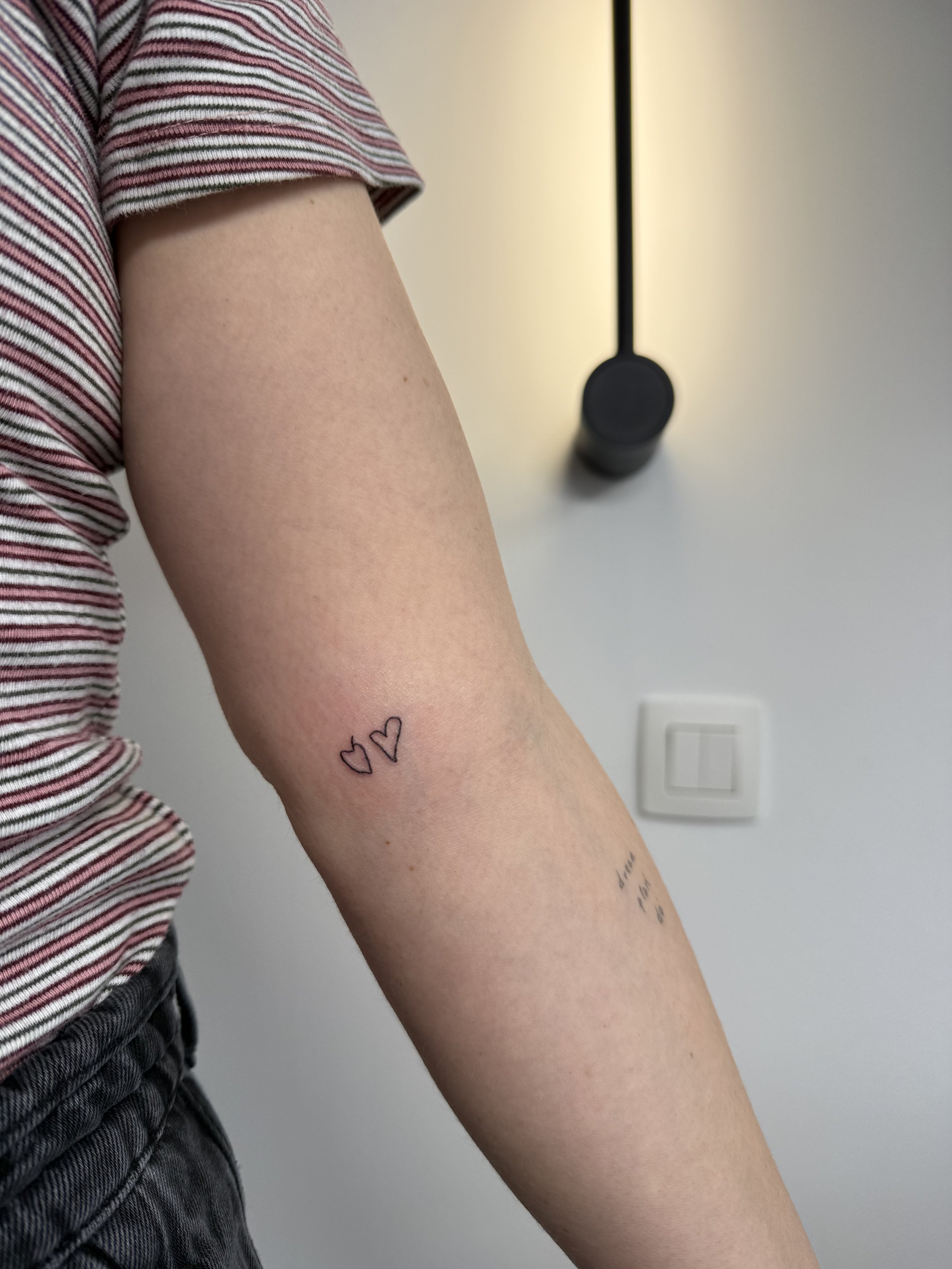 Close-up van een arm met kleine hart-tatoeages en een regenboog-tatoeage, iemand draagt een gestreept shirt en zit in een kamer met een witte muur en een muurcontact.