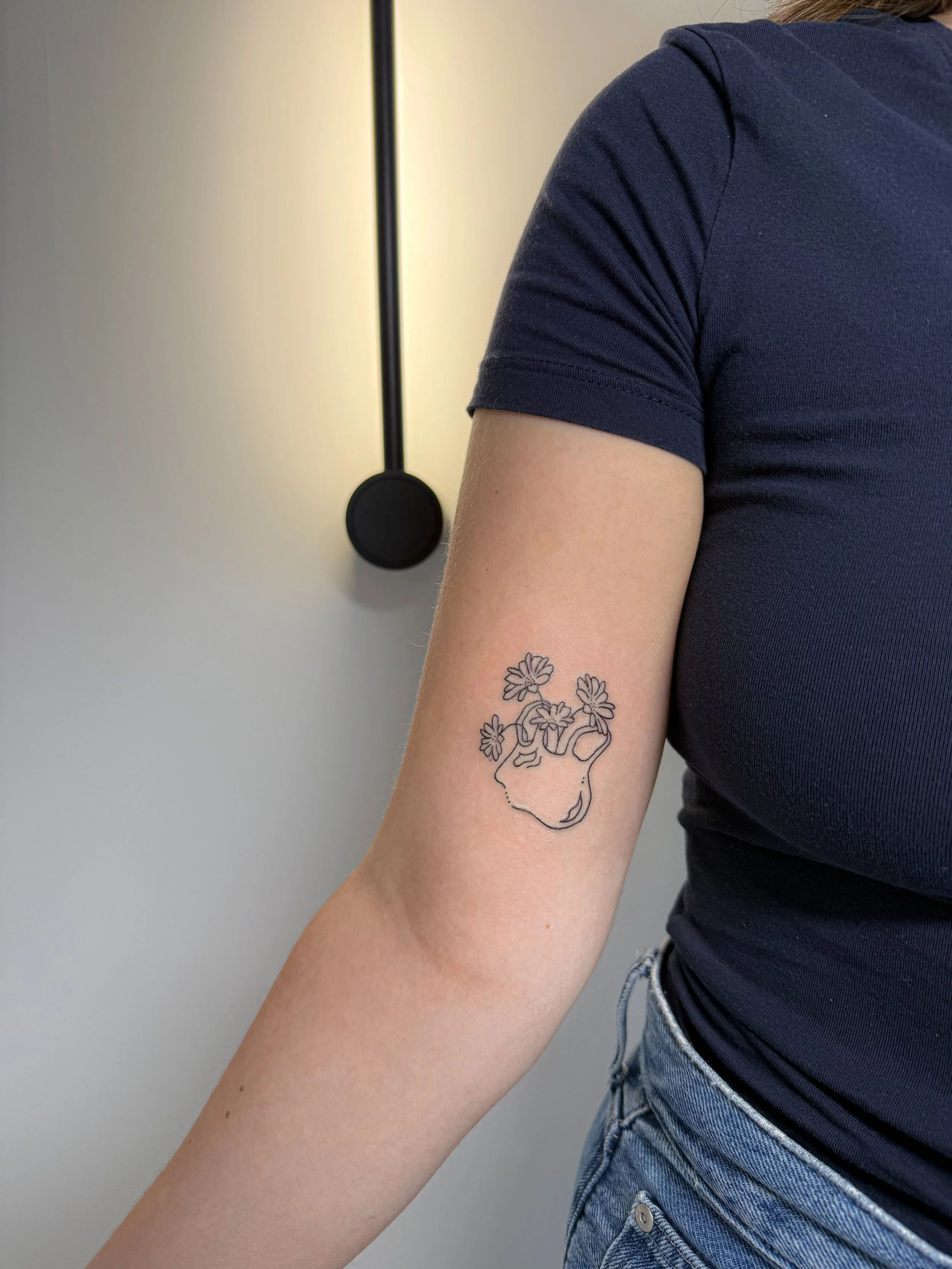 Close-up van een persoon met een tatoeage van een schedel met bloemen op de rechtermouw van een donkerblauw shirt, met een lamp en een muur op de achtergrond.