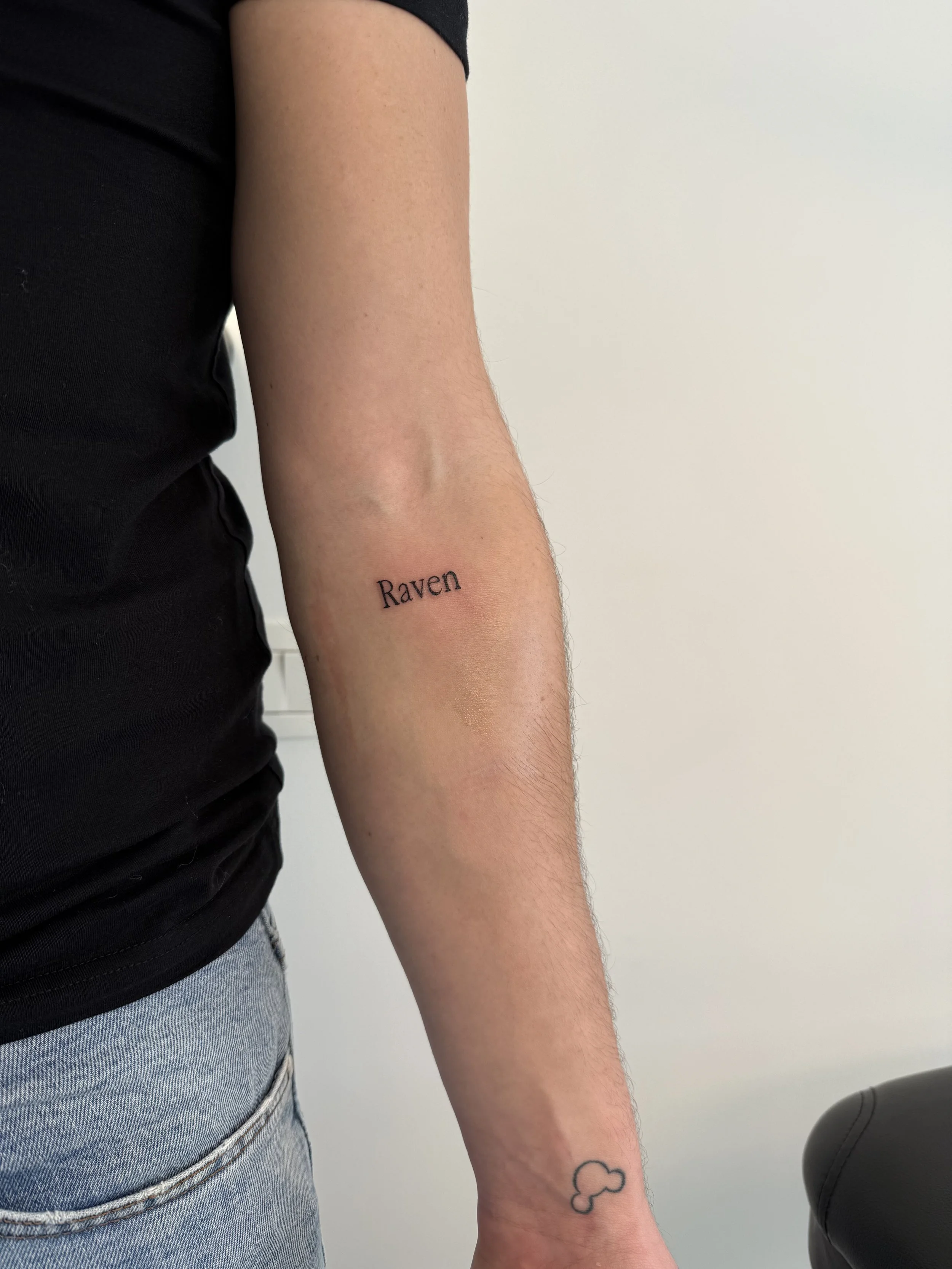 De afbeelding toont een close-up van een rechterarm met een tattoo van een wolk en de tekst 'Raven', gekleed in een zwart shirt, met een witte muur op de achtergrond.