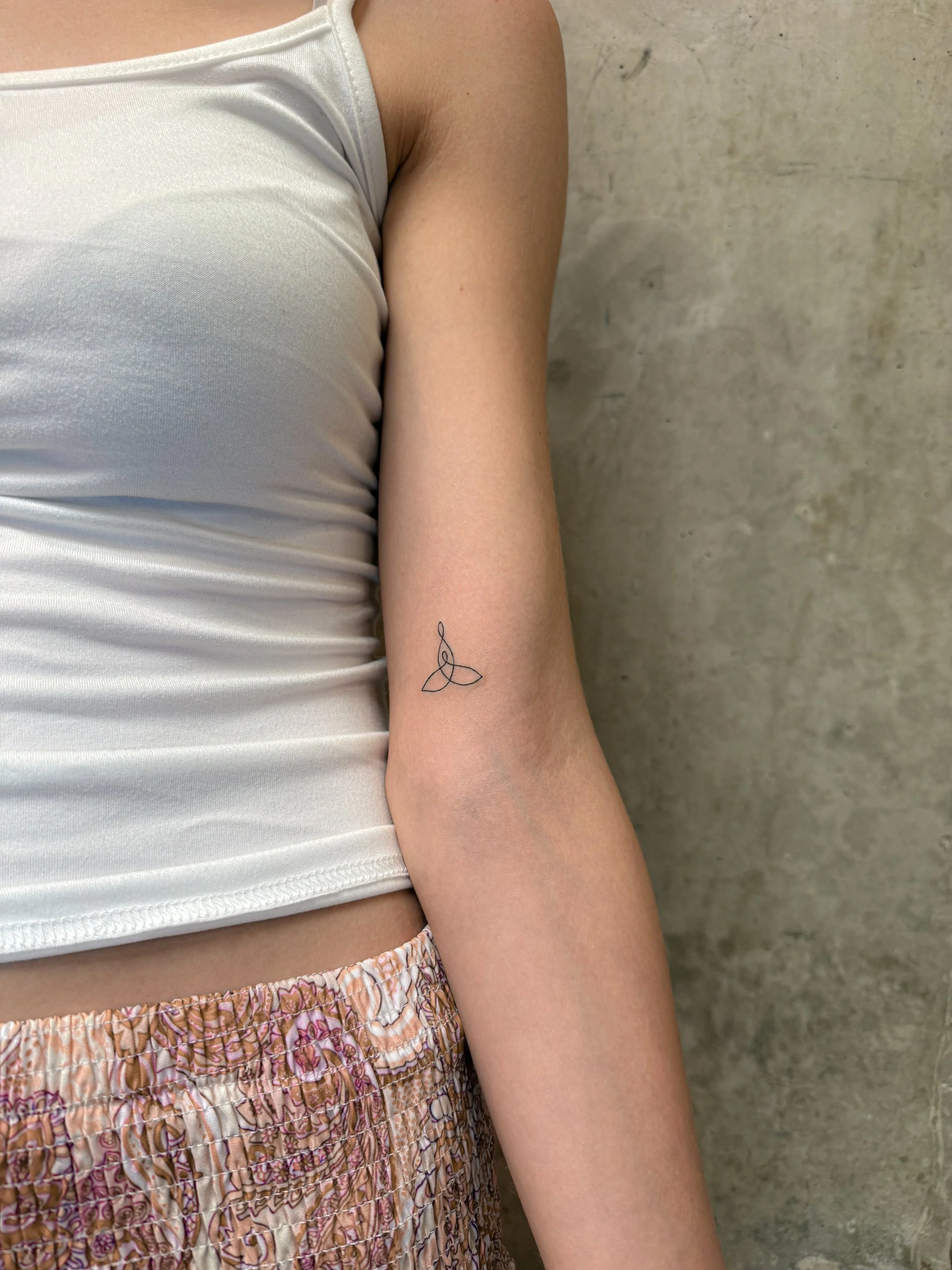 Een close-up van een persoon met een tatoeage op de onderarm, die een geometrisch ontwerp van een drievoudige driehoek bevat, terwijl ze een satijnen top en een geruite rok draagt op een betonnen vloer.