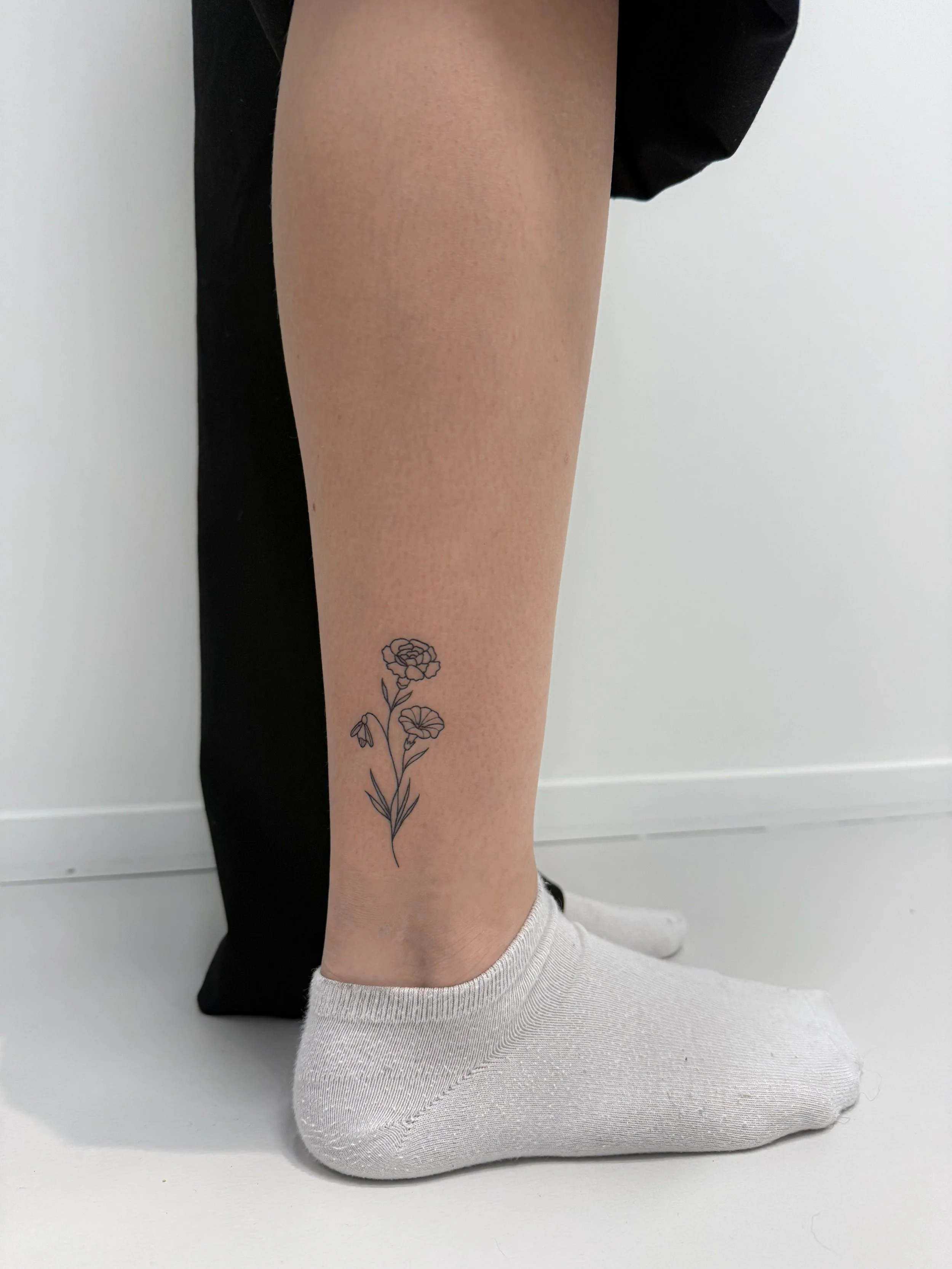 Een onderbeen met een bloemen-tatoeage, gedragen door iemand die witte sokken en een zwarte broek draagt, gekleed in zwarte en witte sneakers.