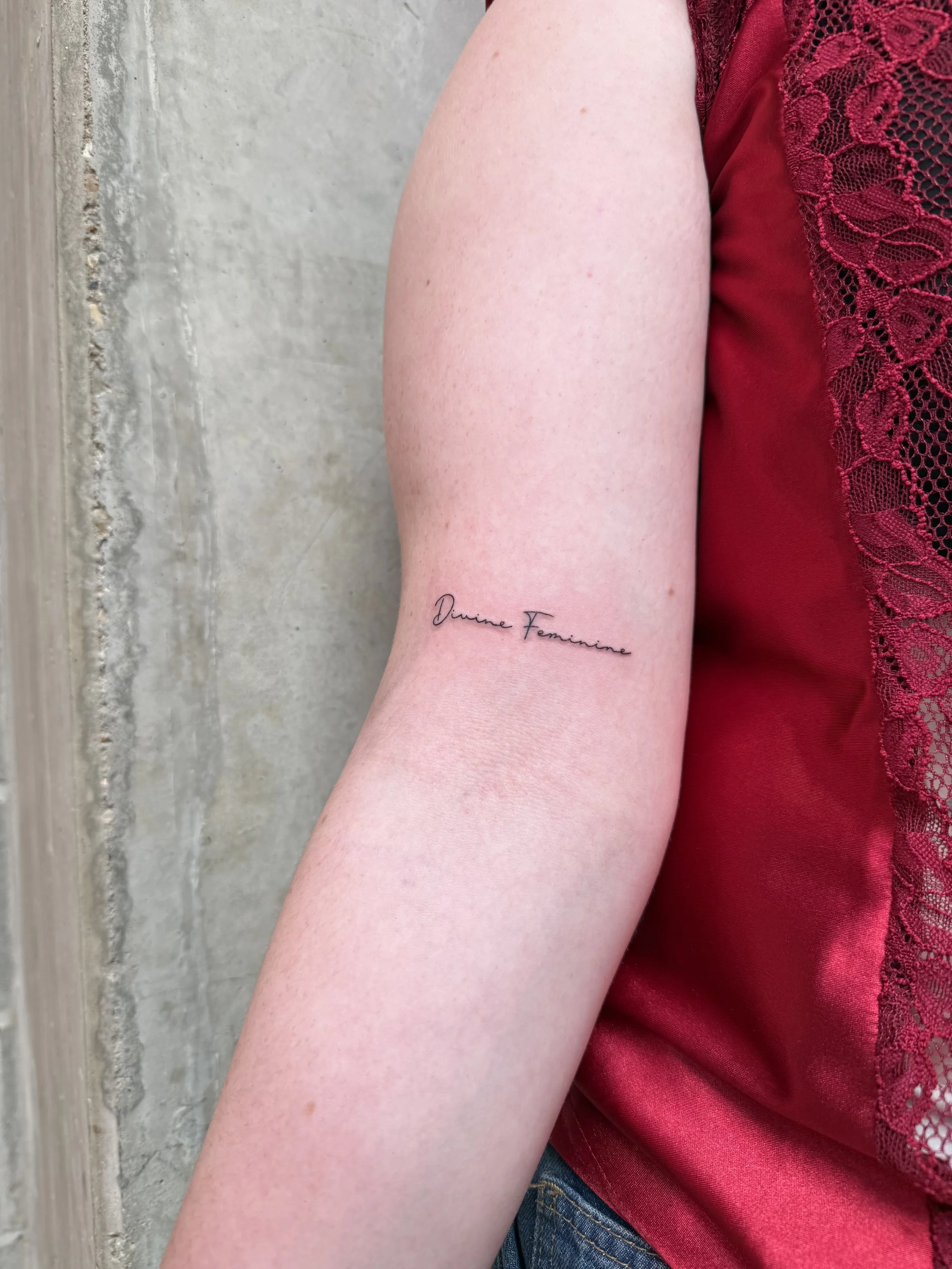 Close-up van een menselijke arm met een tatoeage die zegt 'Divine Feminine', naast een rood kanten shirt.