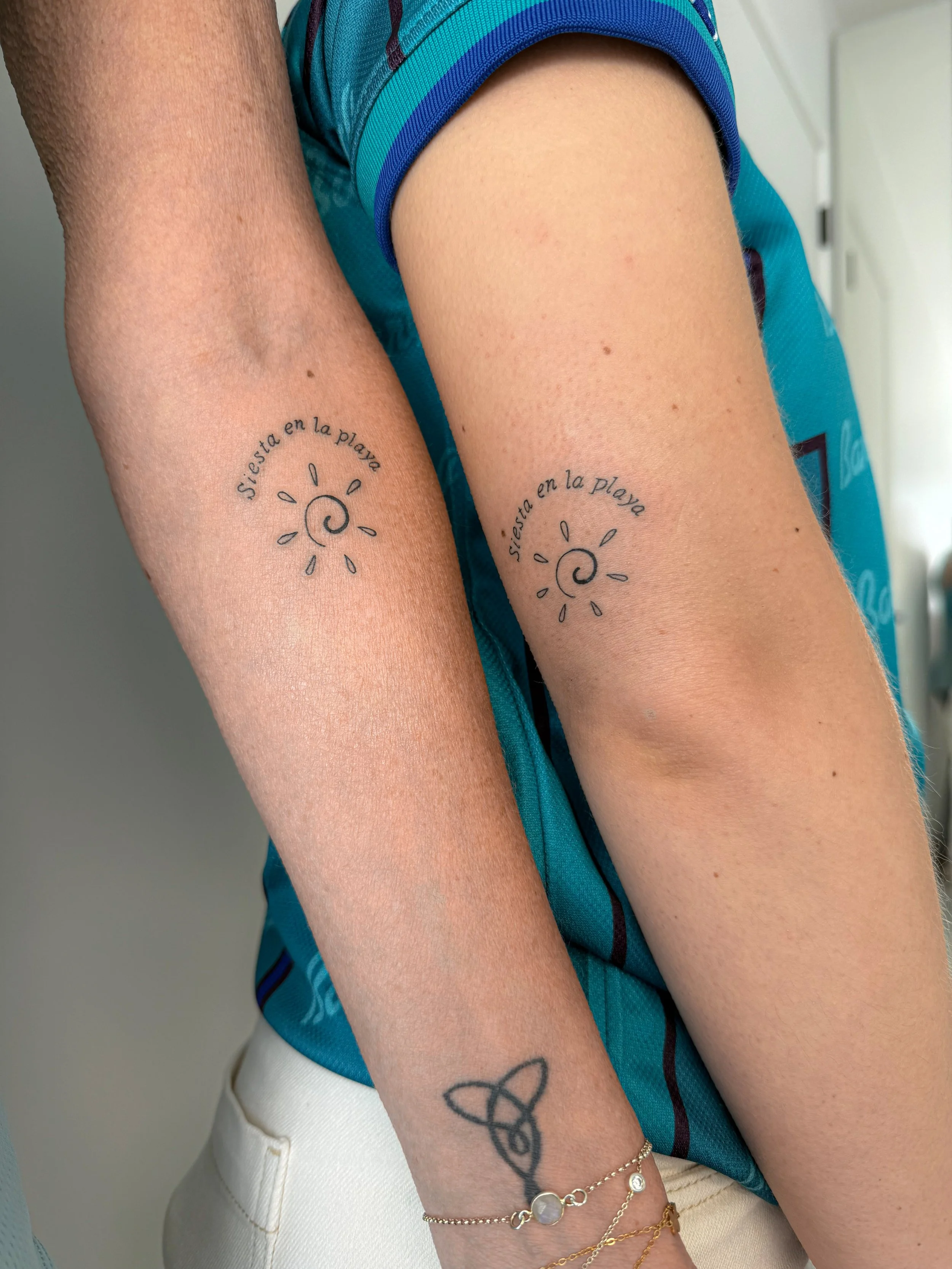 Close-up van twee vrouwenarmen met tattoo's en sieraden, waarvan een met de tekst 'Siesta en la playa' en een spirale, de andere met een geknoopte lijn als tattoo. Ze dragen verschillende geldringen en armbanden.