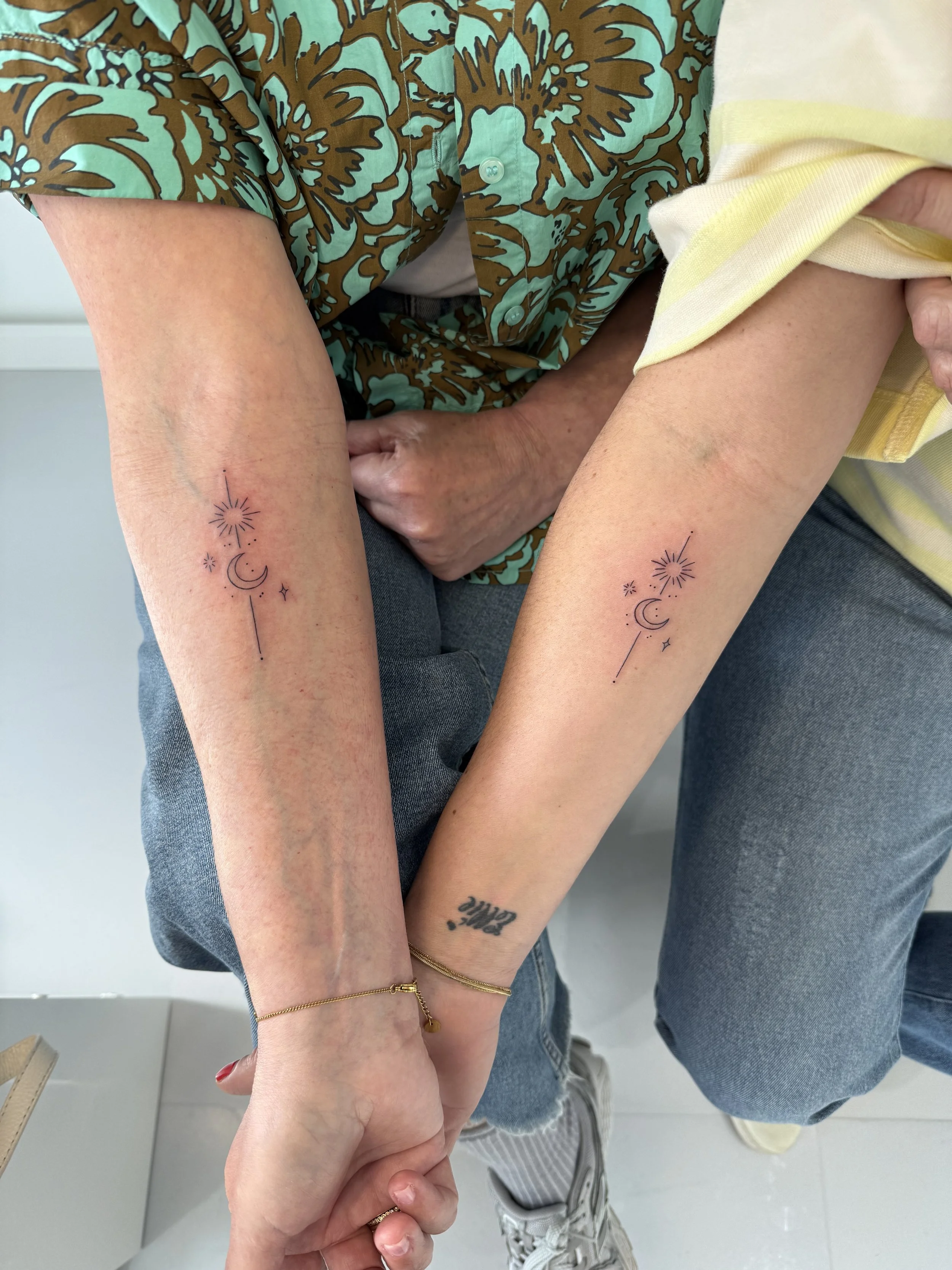 Twee mensen houden elkaars handen vast. Ze hebben beiden tatoeages van een maan, sterren, een zon en lijnen op hun onderarmen.