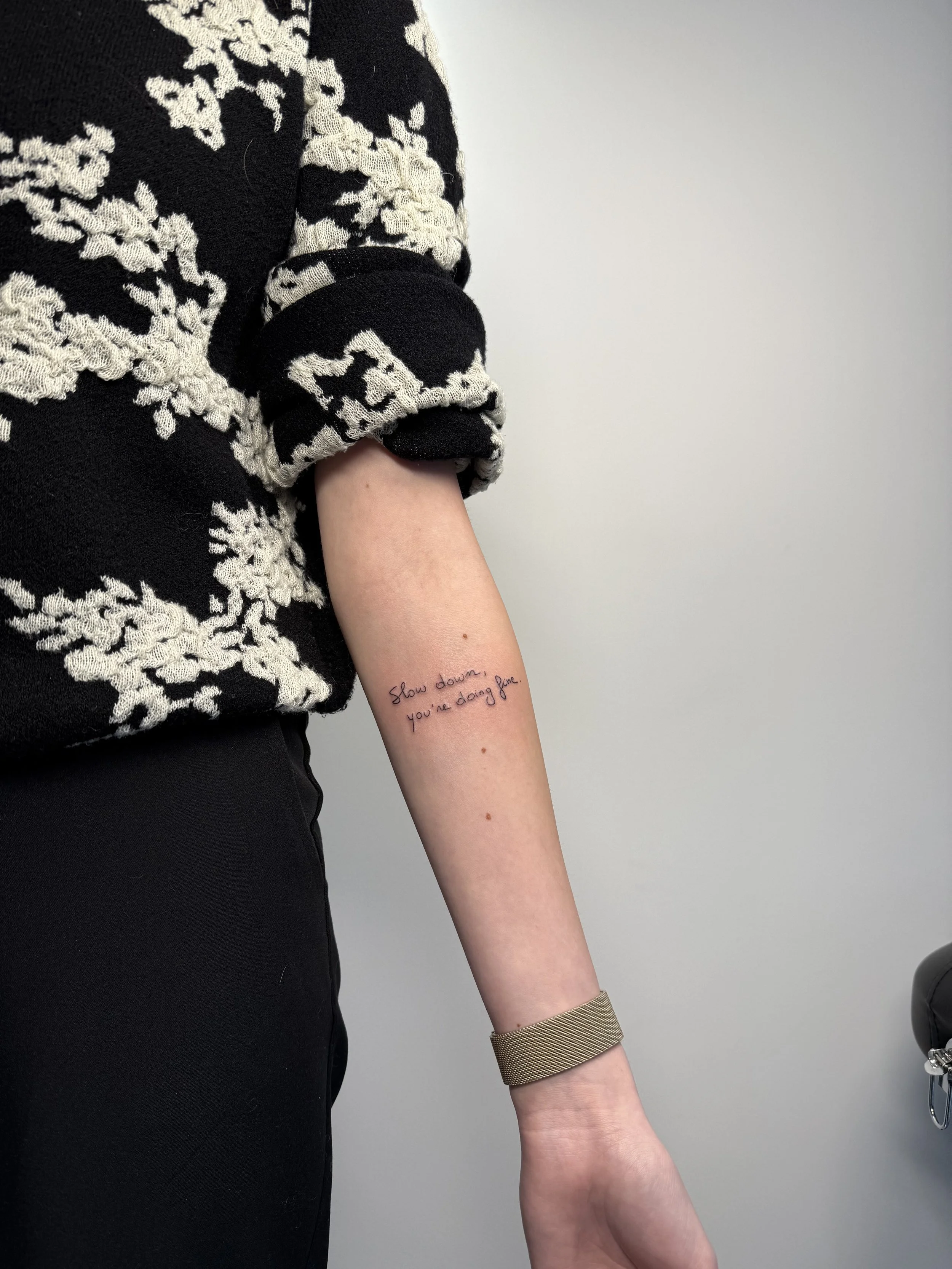 Een close-up van een arm met een tattoo die zegt: 'Slow down, you're doing fine' en een manchette armband, naast een persoon die een zwart-wit knielange jurk draagt.
