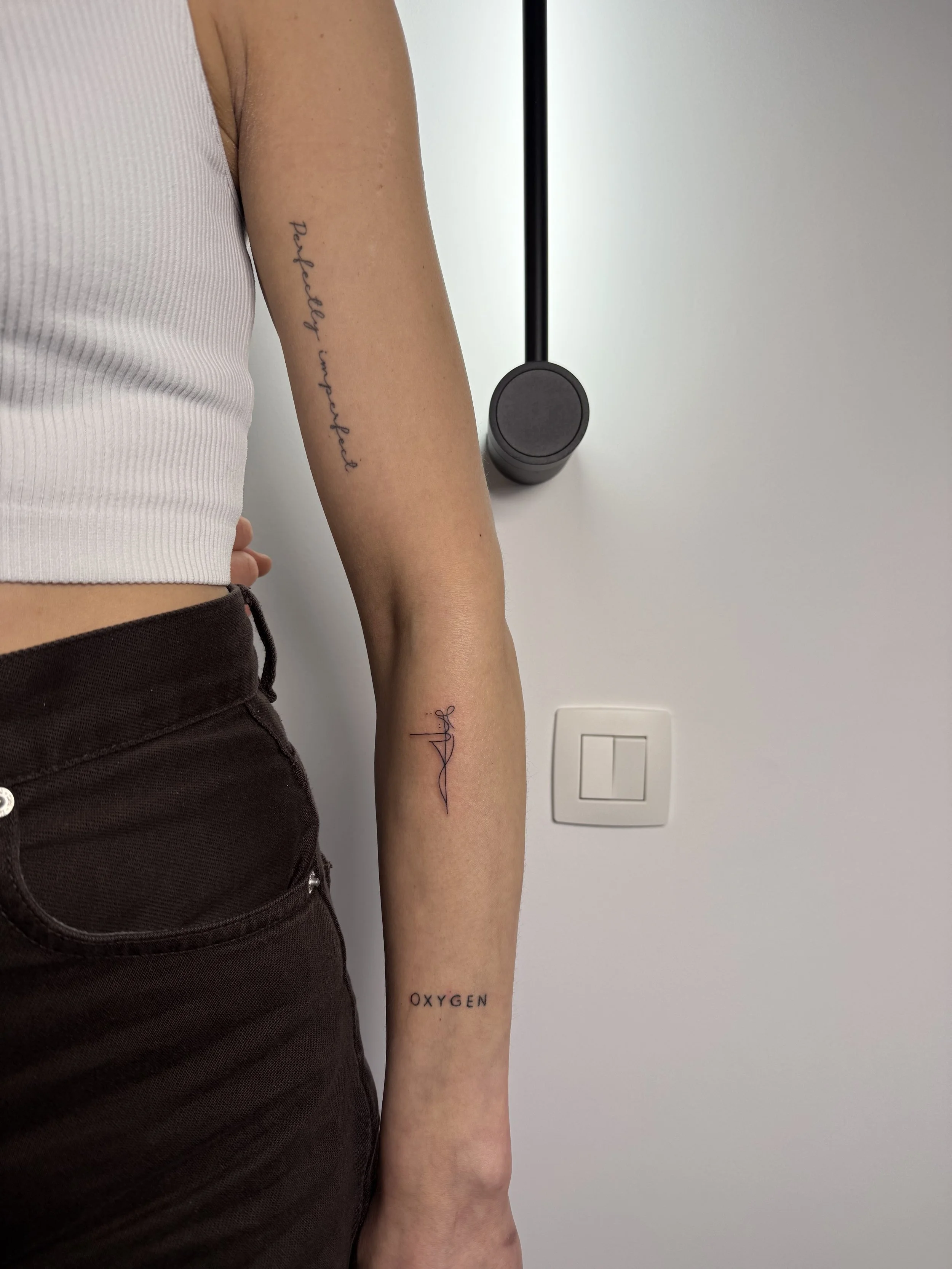 De arm van een persoon met tijdelijke tattoo's, met woorden zoals 'OXYGEN', 'Peacefully imperfect' en een streepje met een handtekening, tegen een witte achtergrond.