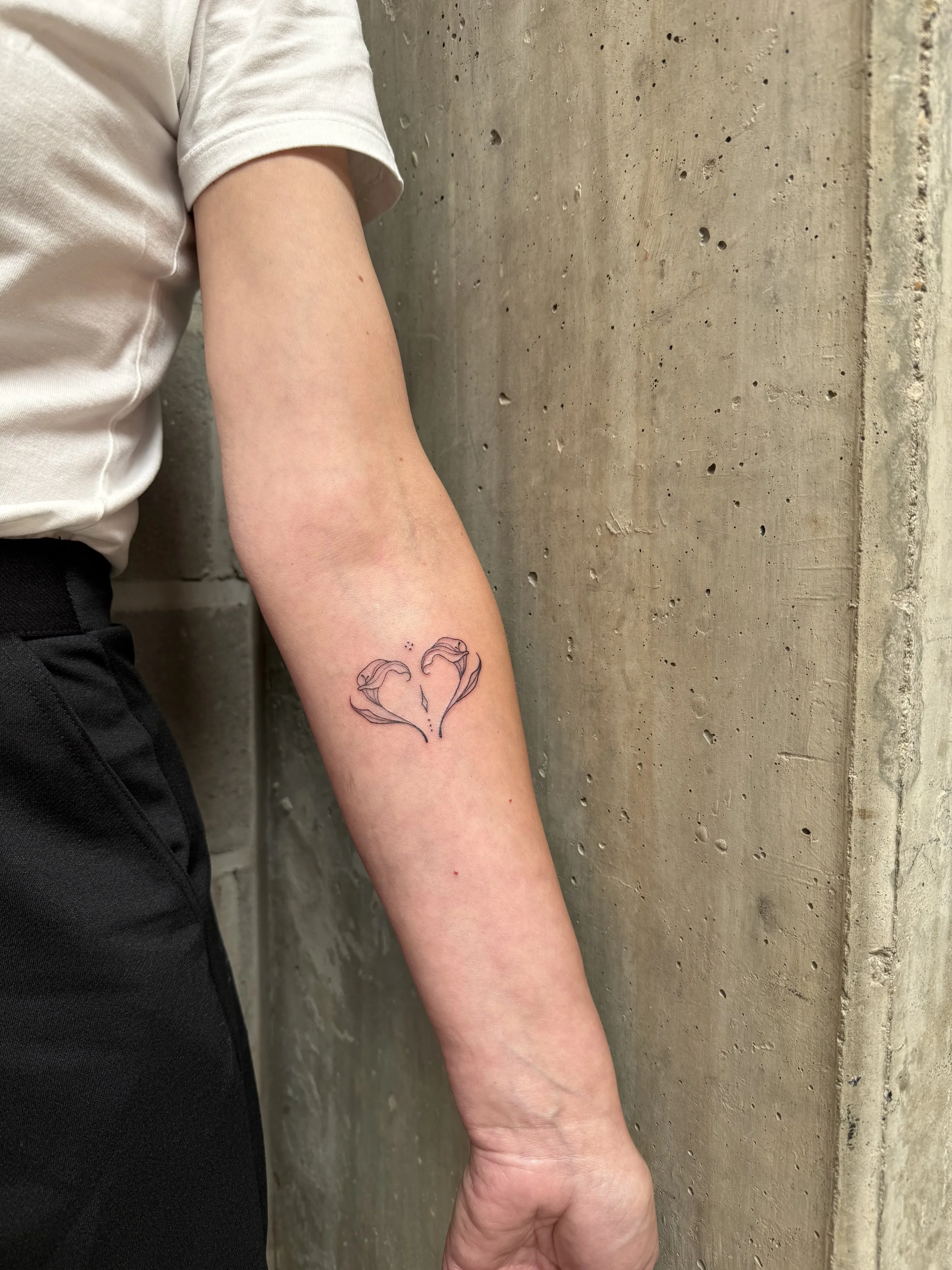 Tienerkind met een tattoo van twee vrouwenhoofden die een hart vormen op de onderarm, gekleed in een wit T-shirt en zwarte broek, leunend tegen een betonnen muur.