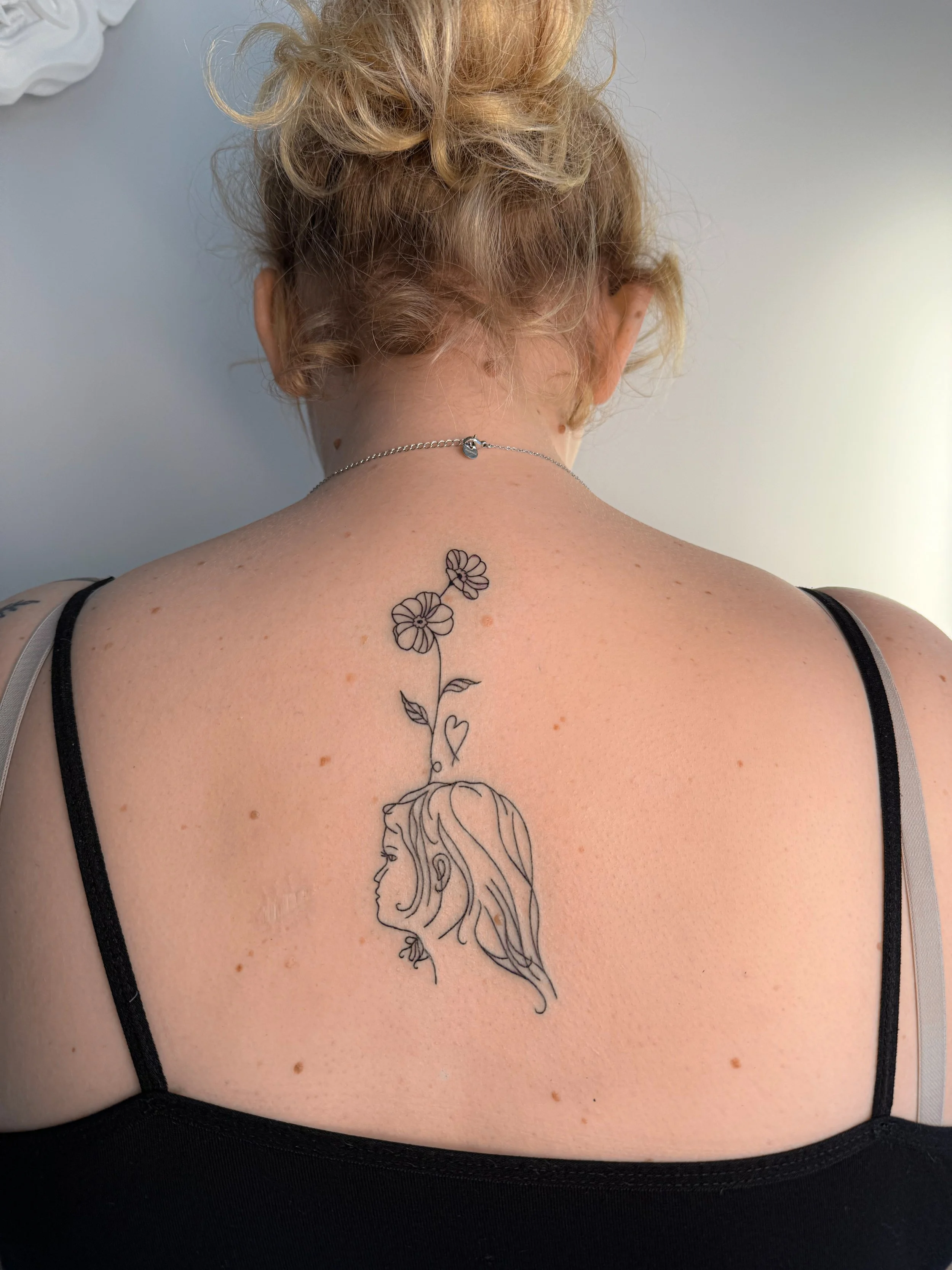 De afbeelding toont de achterkant van een vrouw met een tattoo van een meisjesprofiel met lang haar en bloemen op haar rug, ze draagt een zwart kanten kledingstuk.