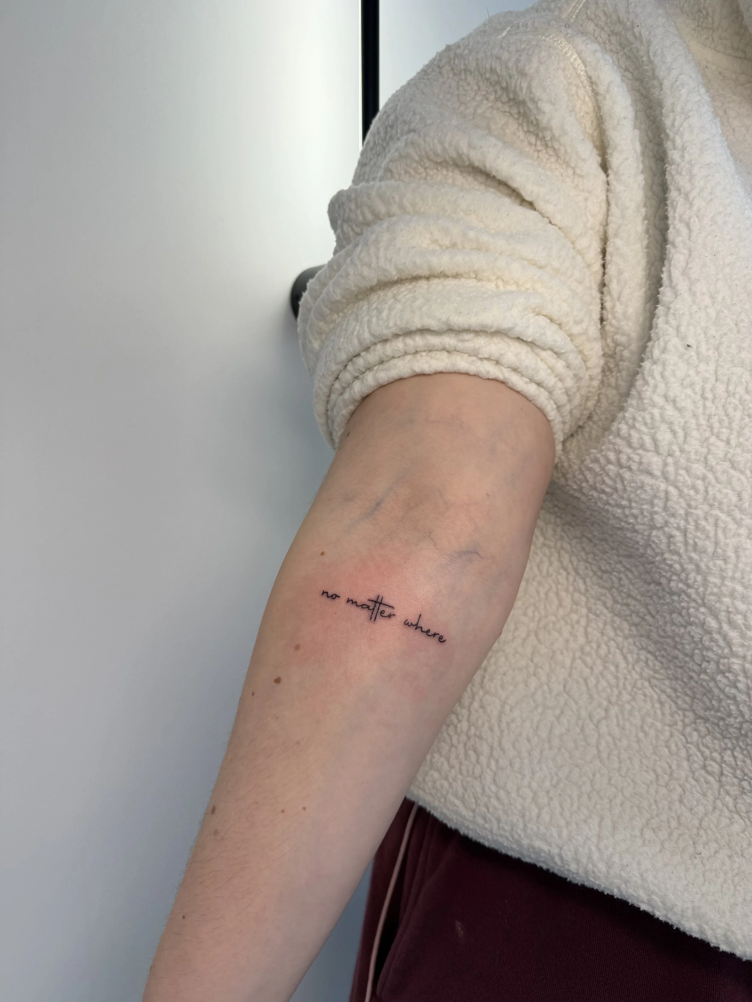 Close-up van een arm met een tatoeage die zegt 'no matter where' en een persoon draagt een beige, zachte fleece jas en een donkerrode broek.