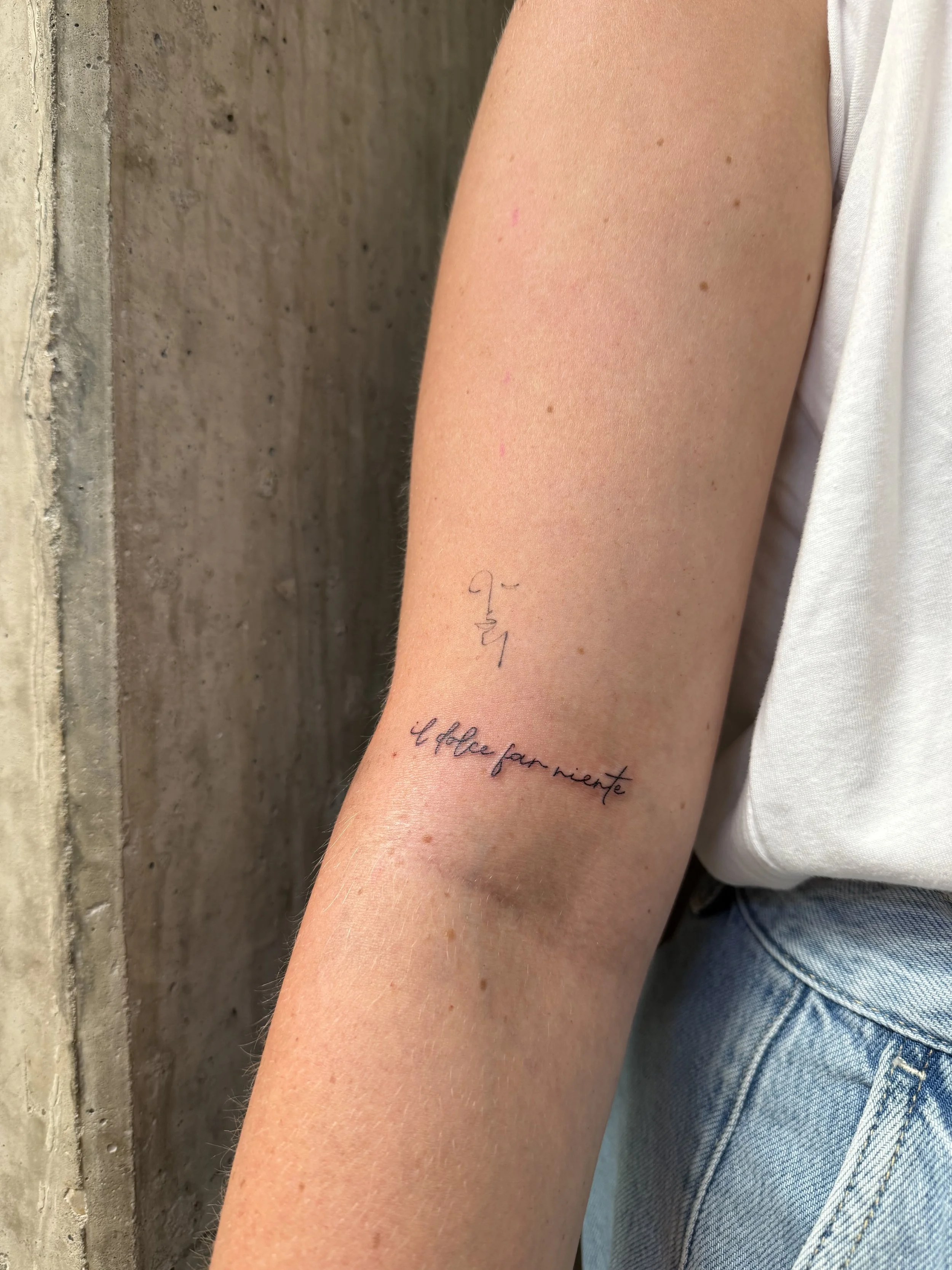 Een close-up van een onderarm met een tatoeage in het zwart dat zegt 'life' en 'coffee for night'. Achtergrond van een betonnen muur, iemand draagt een witte T-shirt en blauwe jeans.
