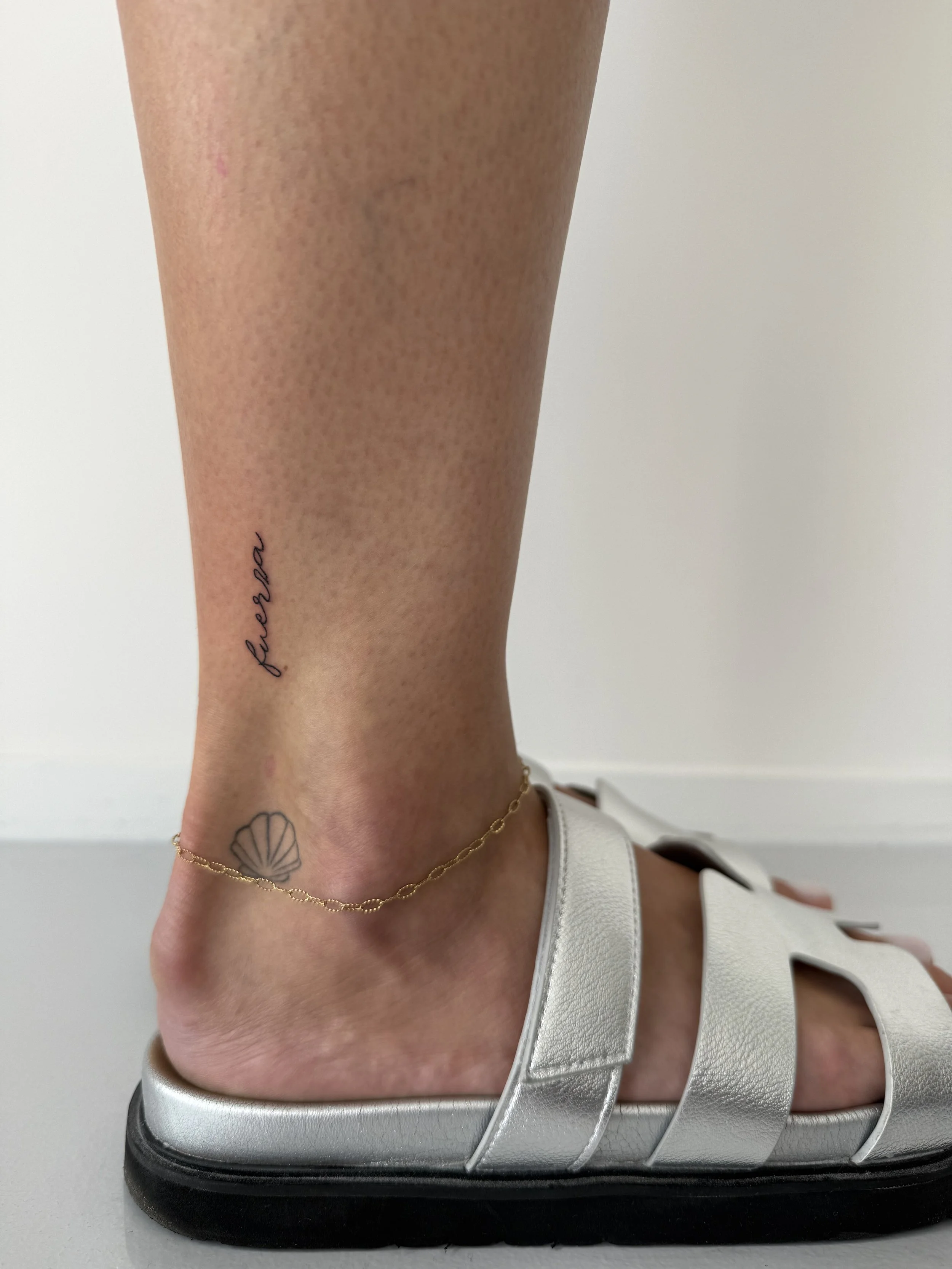 Een voet in een witte sandal met banden, een gouden enkelketting en een tattoo van een schelp op de enkel, geplaatst op een sportschoen.