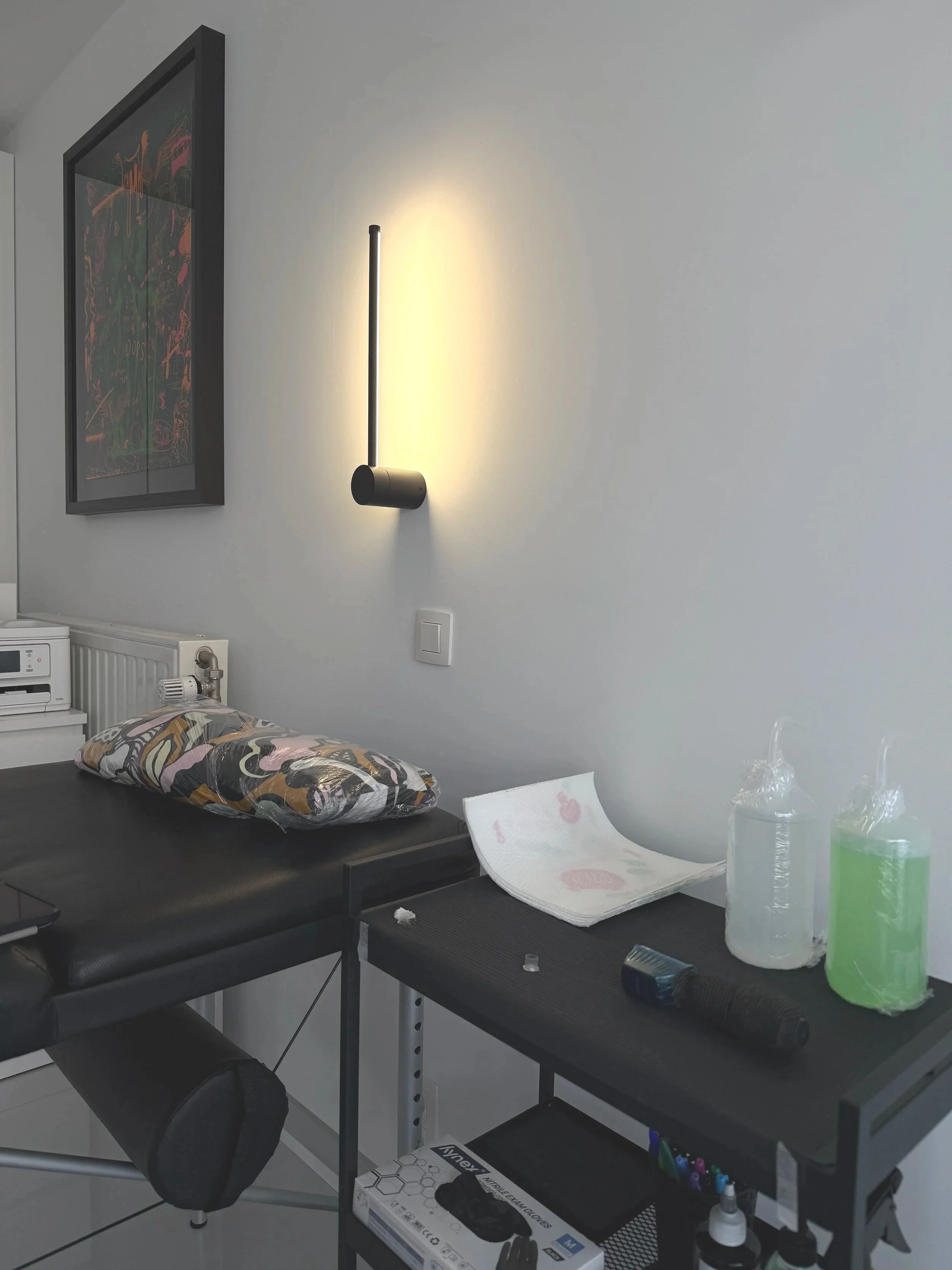 Een tafel met medische benodigdheden zoals een plastic cup, een doek, en een geluidloos instrument. Er hangt een lamp aan de muur en er is een schilderij. De ruimte lijkt een medische of laboratoriumomgeving.